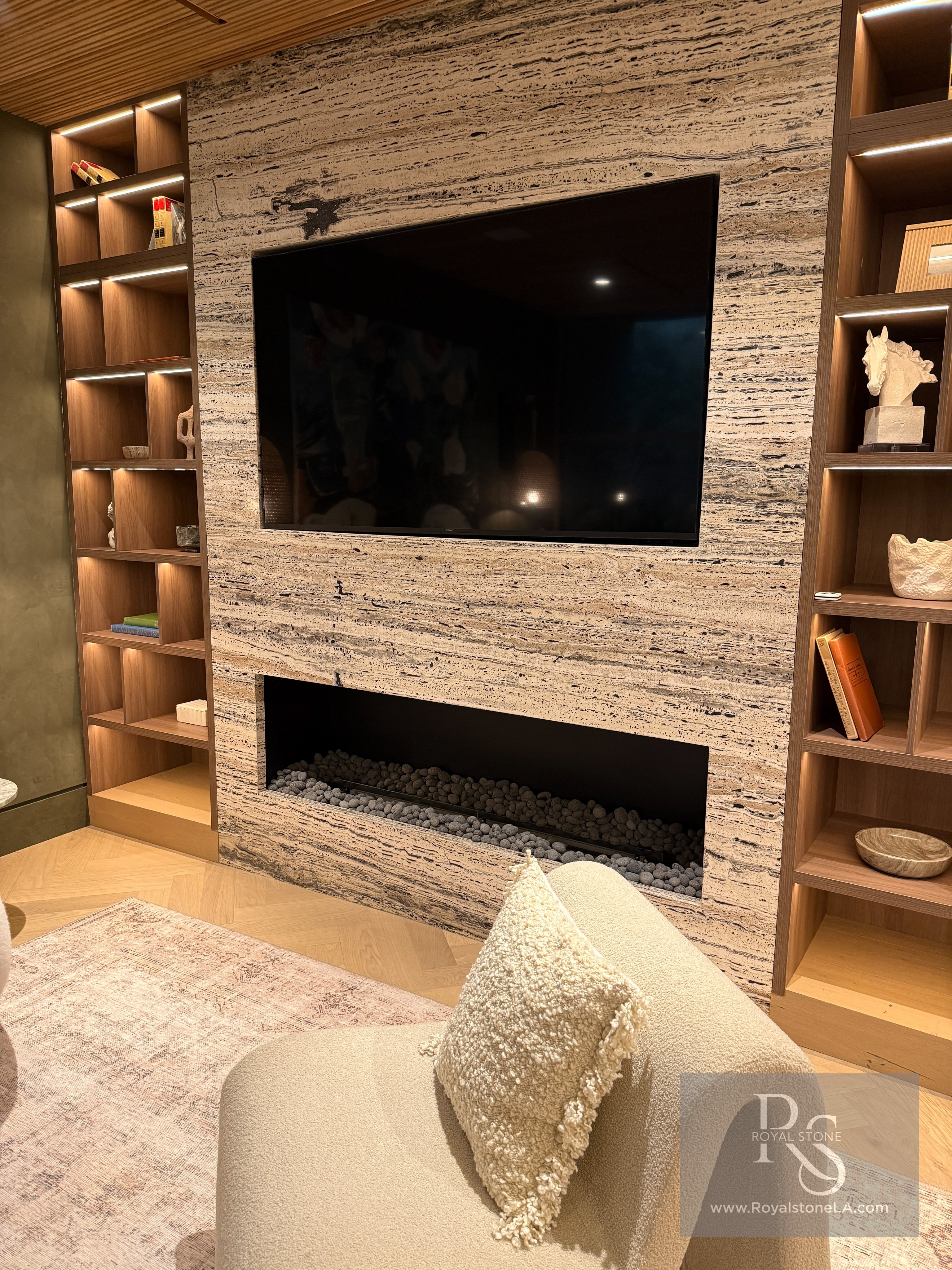 Antolini Black Label Platinum Travertine vein cut and leathered installed as a full-height fireplace and media wall, with a wall-mounted Sony display, a linear gas hearth filled with grey lava stones, walnut library millwork flanking the wall, and a herringbone oak floor