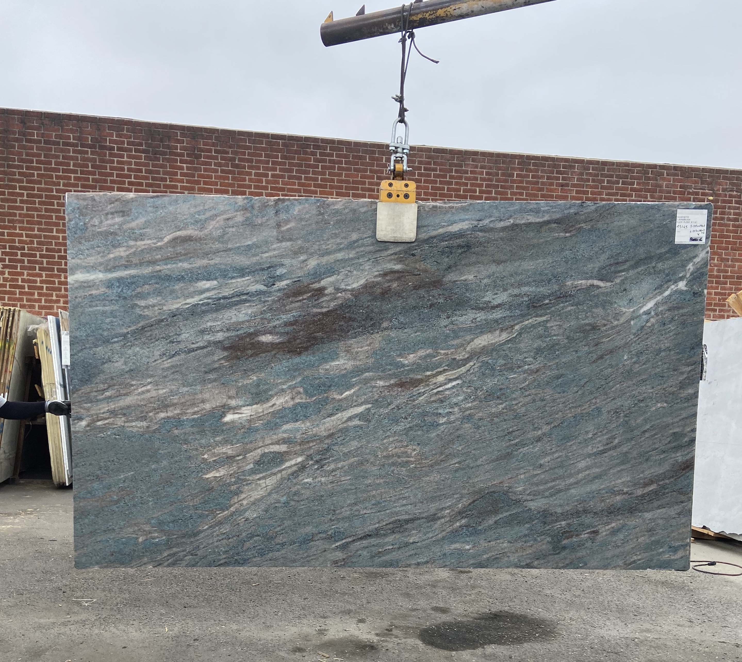 Antolini Caribbean Blue quartzite slab, 79 by 134 inches, polished finish with soft sea-blue field swept by diagonal currents of cream, rust, and burgundy, lifted by crane against a brick wall at Royal Stone in Los Angeles