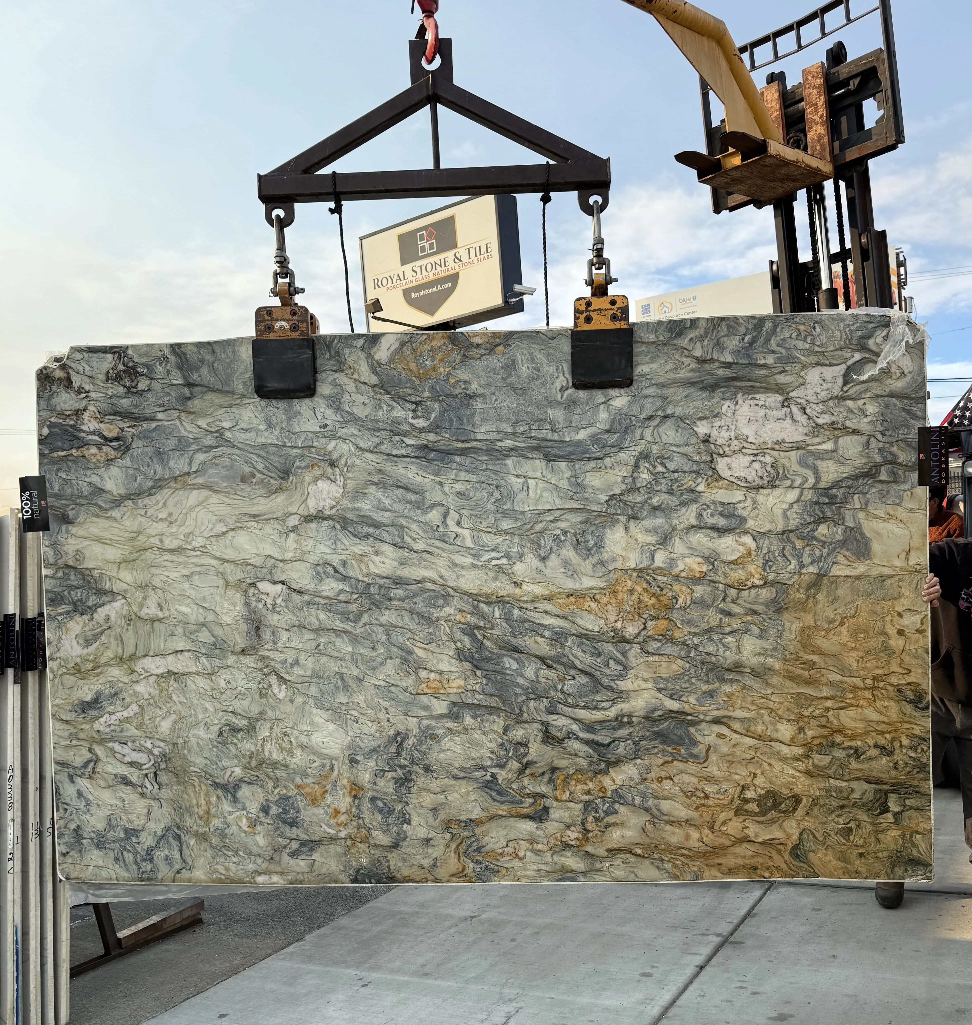 Antolini Fusion quartzite slab on the crane in front of the Royal Stone & Tile sign, pale sage and gray field with amber and bronze veining