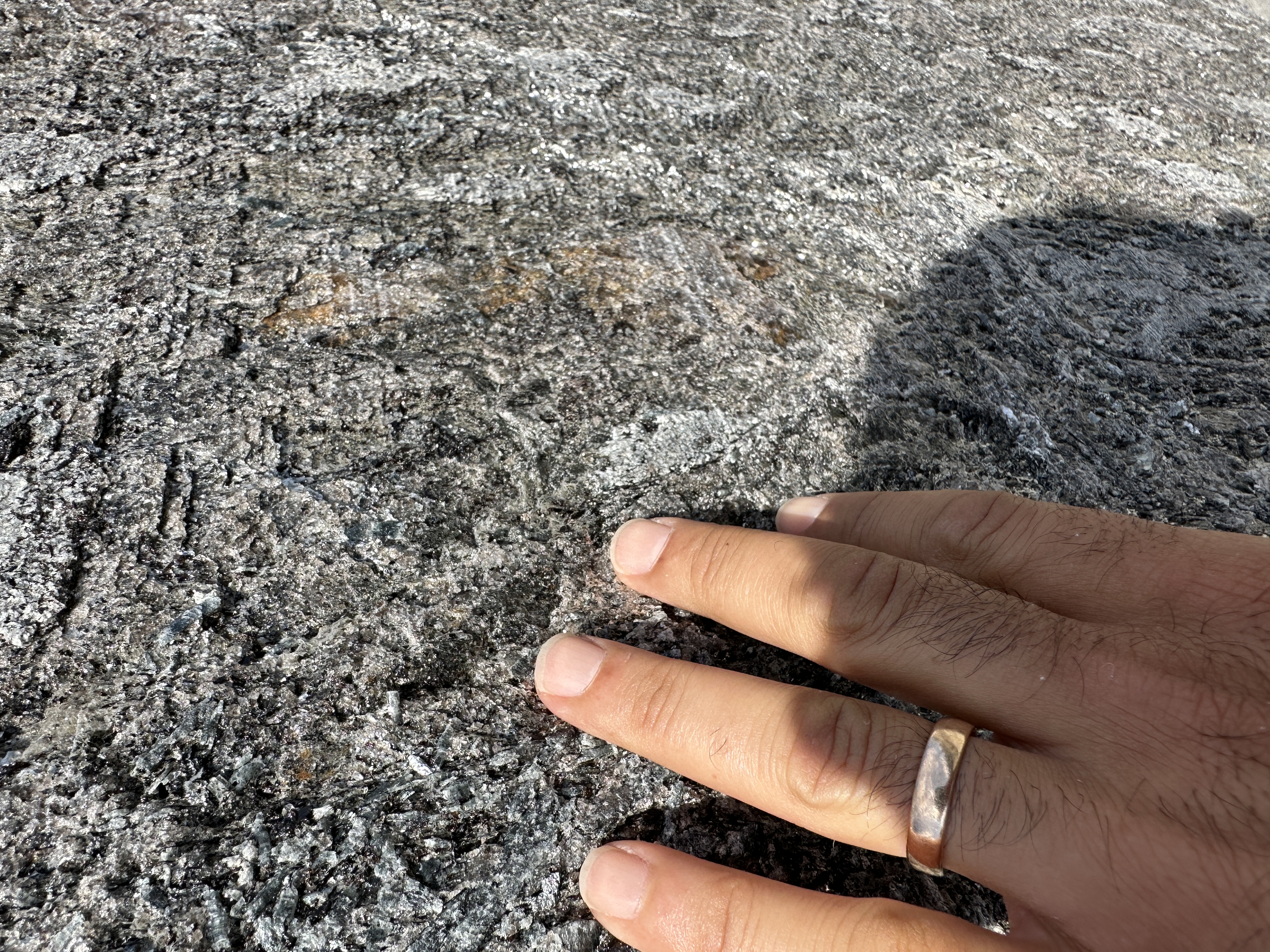 Close-up of Antolini Kosmos Quartzite Super Cleft Texture finish with a hand for scale, showing the rough split rock face, silver mica flecks, charcoal ground, and rust-coloured iron passages