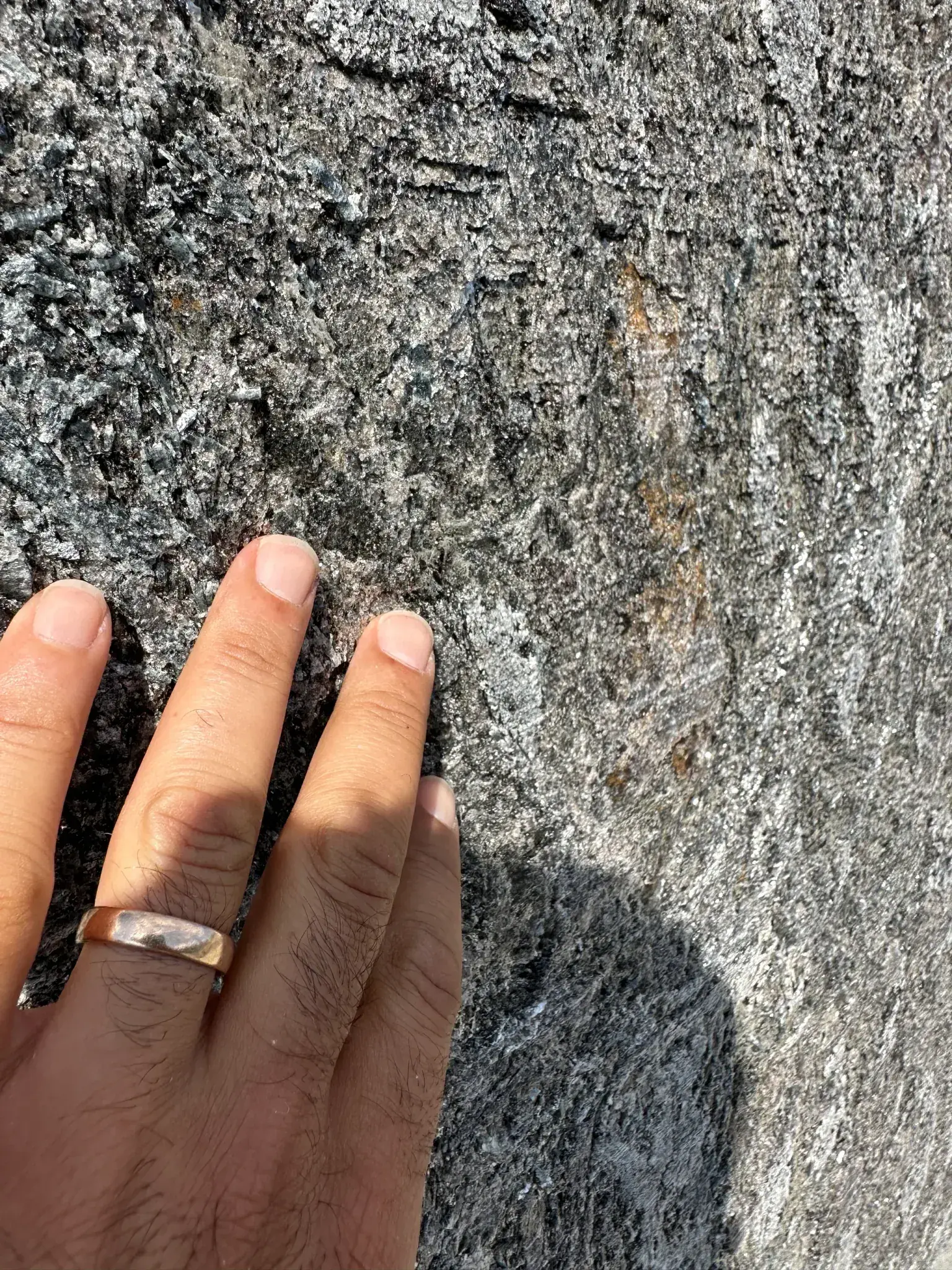 Close-up of Antolini Kosmos Quartzite Super Cleft Texture finish with a hand for scale, showing the rough split rock face, silver mica flecks, charcoal ground, and rust-coloured iron passages