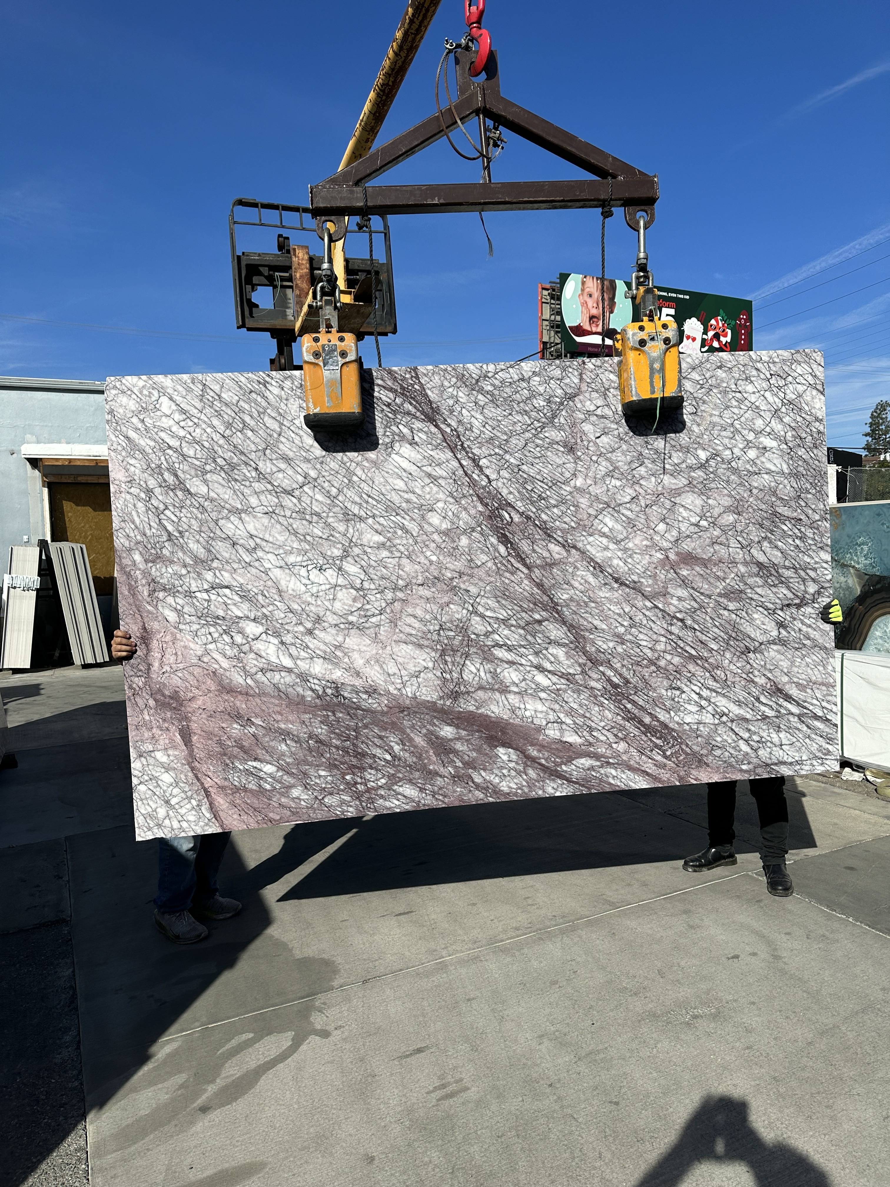 Antolini Lilac Viola Premium Extra Select marble slab, 103 by 63 inches, polished finish, dusty rose and lilac field webbed with a dense net of fine plum and near-black veining with pockets of brighter pink and white quartz, lifted by crane under blue sky at Royal Stone in Los Angeles