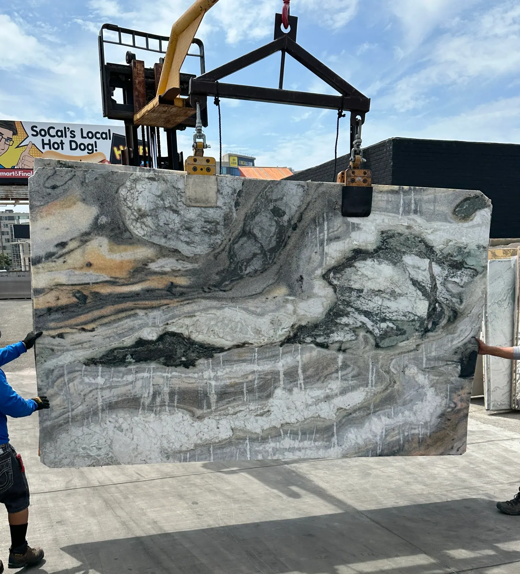 2cm Antolini Midnight Ocean Wave marble slab, 78 by 125 inches, honed finish, pictorial composition of cresting charcoal and gunmetal waves with amber and burnt-orange passages above white quartz spray and vertical rain-like streaks below, lifted by crane against a partly cloudy sky at Royal Stone in Los Angeles