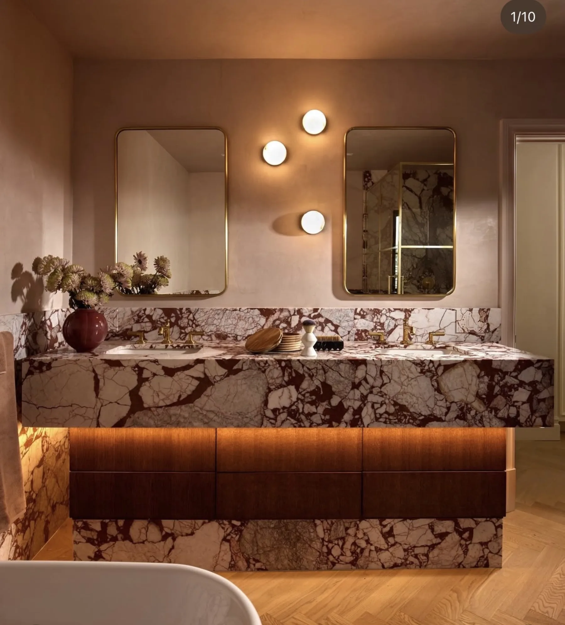Double vanity wrapped in Calacatta Viola Rosso Extra marble — bookmatched apron and deck against plaster walls, brass fittings, dark wood drawer fronts