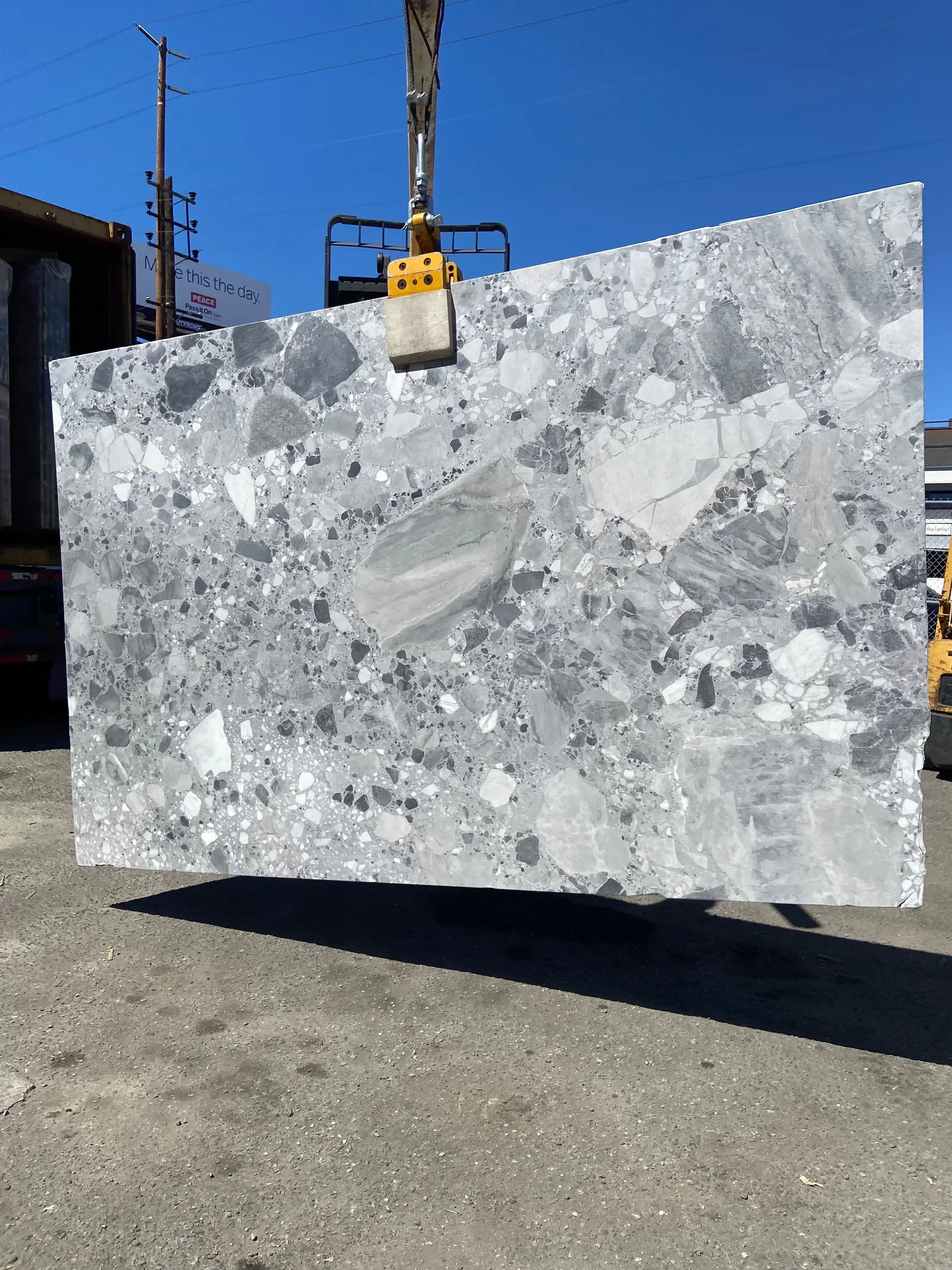 Ceppo Grigio Oreo marble slab, honed, a cool grey conglomerate crowded with bold black-and-white clasts, suspended from a crane clamp in the Royal Stone yard in Los Angeles under a clear blue sky