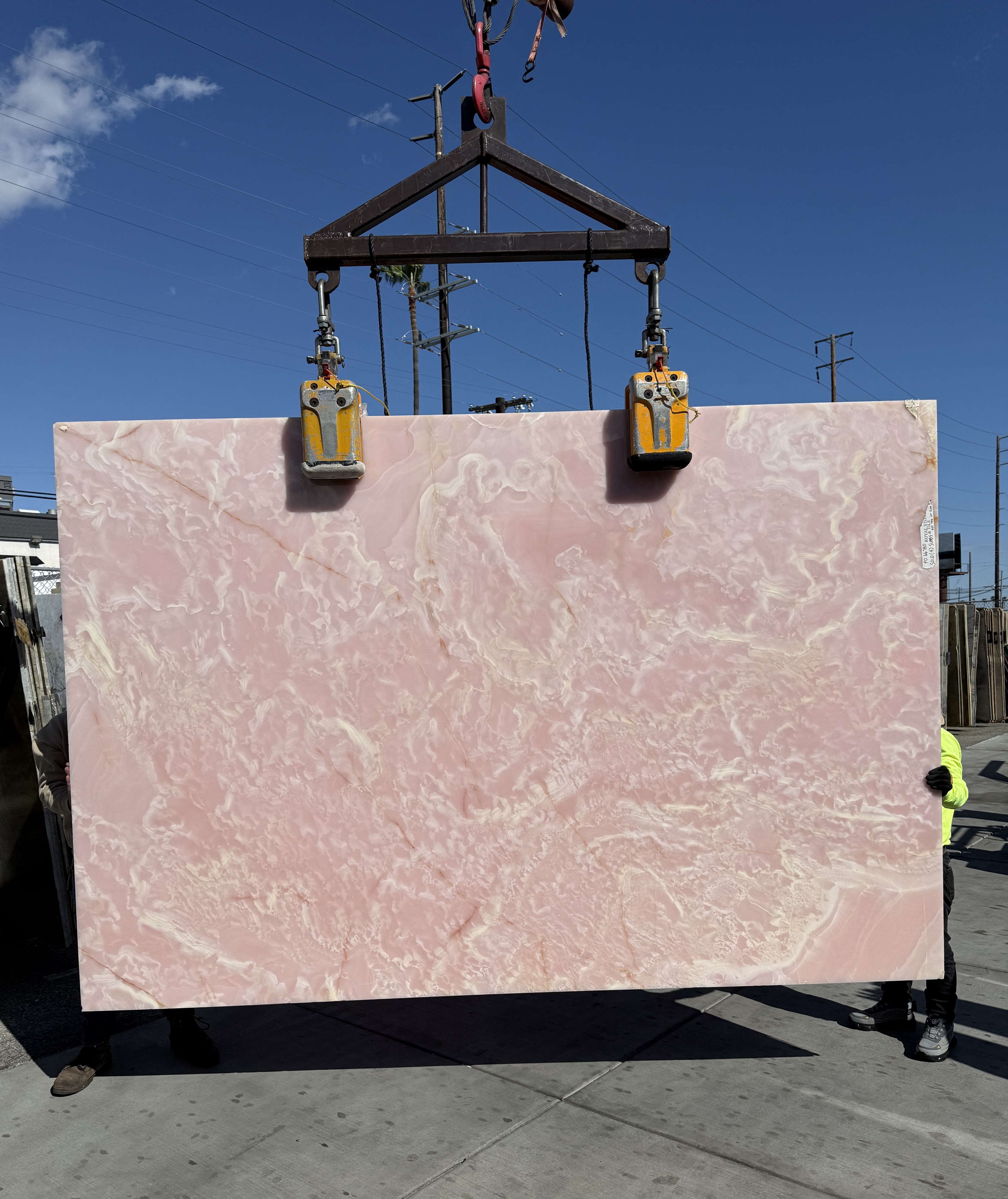Pink Onyx Onice Rosa polished 1.5cm slab lifted on the crane against blue sky at Royal Stone Los Angeles, saturated blush body with translucent cream and white drift