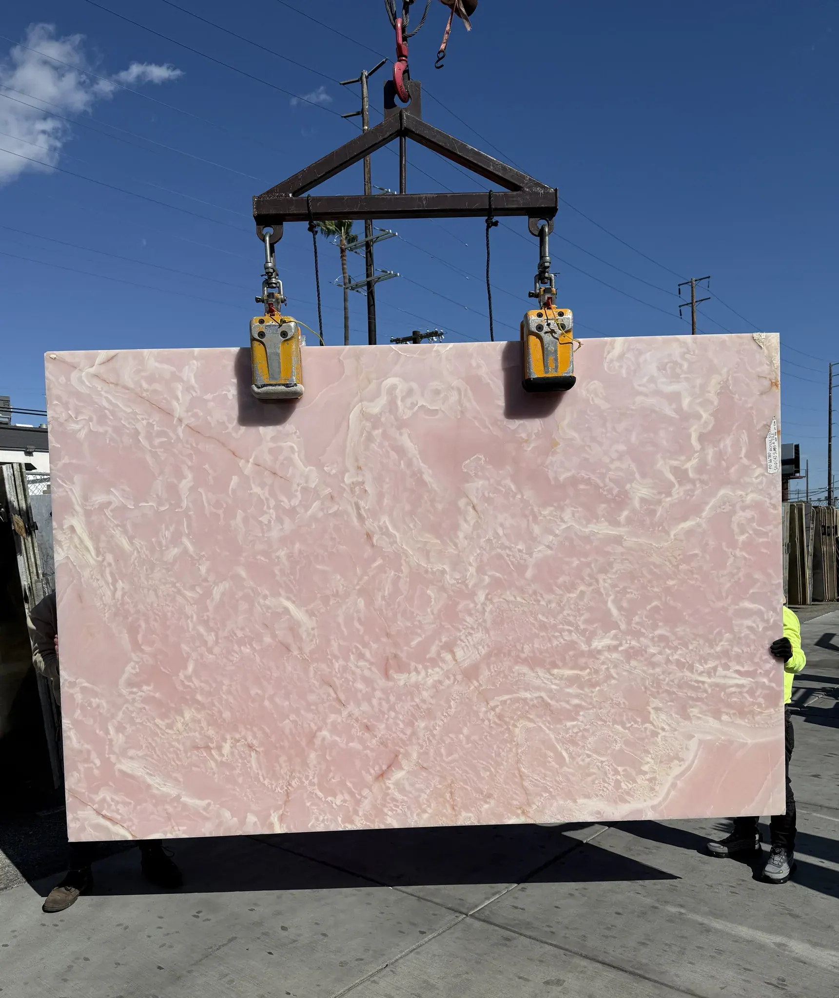 Pink Onyx Onice Rosa polished 1.5cm slab lifted on the crane against blue sky at Royal Stone Los Angeles, saturated blush body with translucent cream and white drift