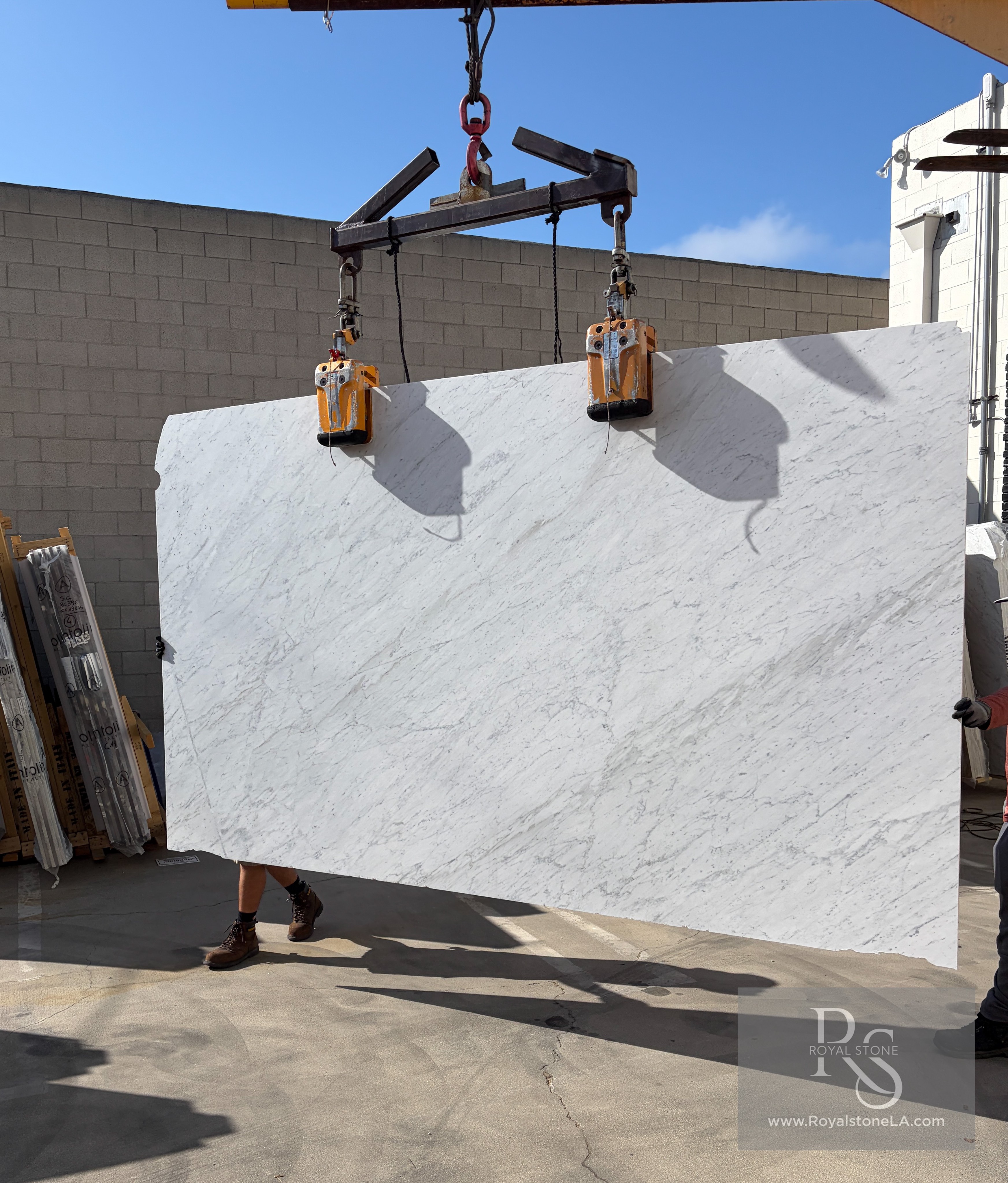 2cm Super Carrara White Select Petris honed marble slab, 75 by 129 inches, bright chalk-white field with soft feathered grey veining, lifted on vacuum suction cups under daylight at the Royal Stone Los Angeles yard