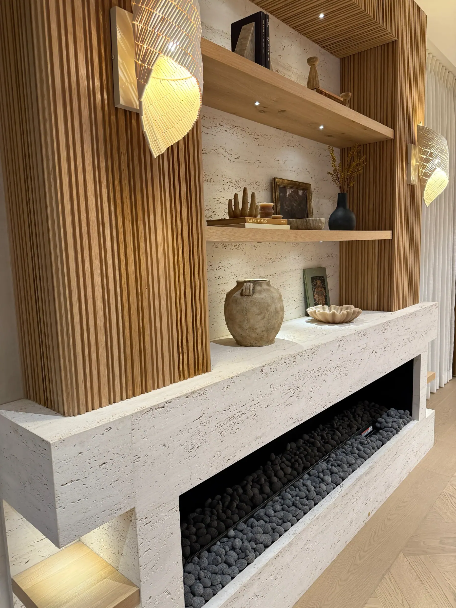 Travertino Sandblasted Cemento vein cut ivory travertine installed as a wrapped fireplace mass with a long horizontal hearth filled with grey lava pebbles, flanked by fluted white oak millwork and a vertical back niche of the same travertine, with floating white oak shelving, woven pendant sconces, and a herringbone oak floor