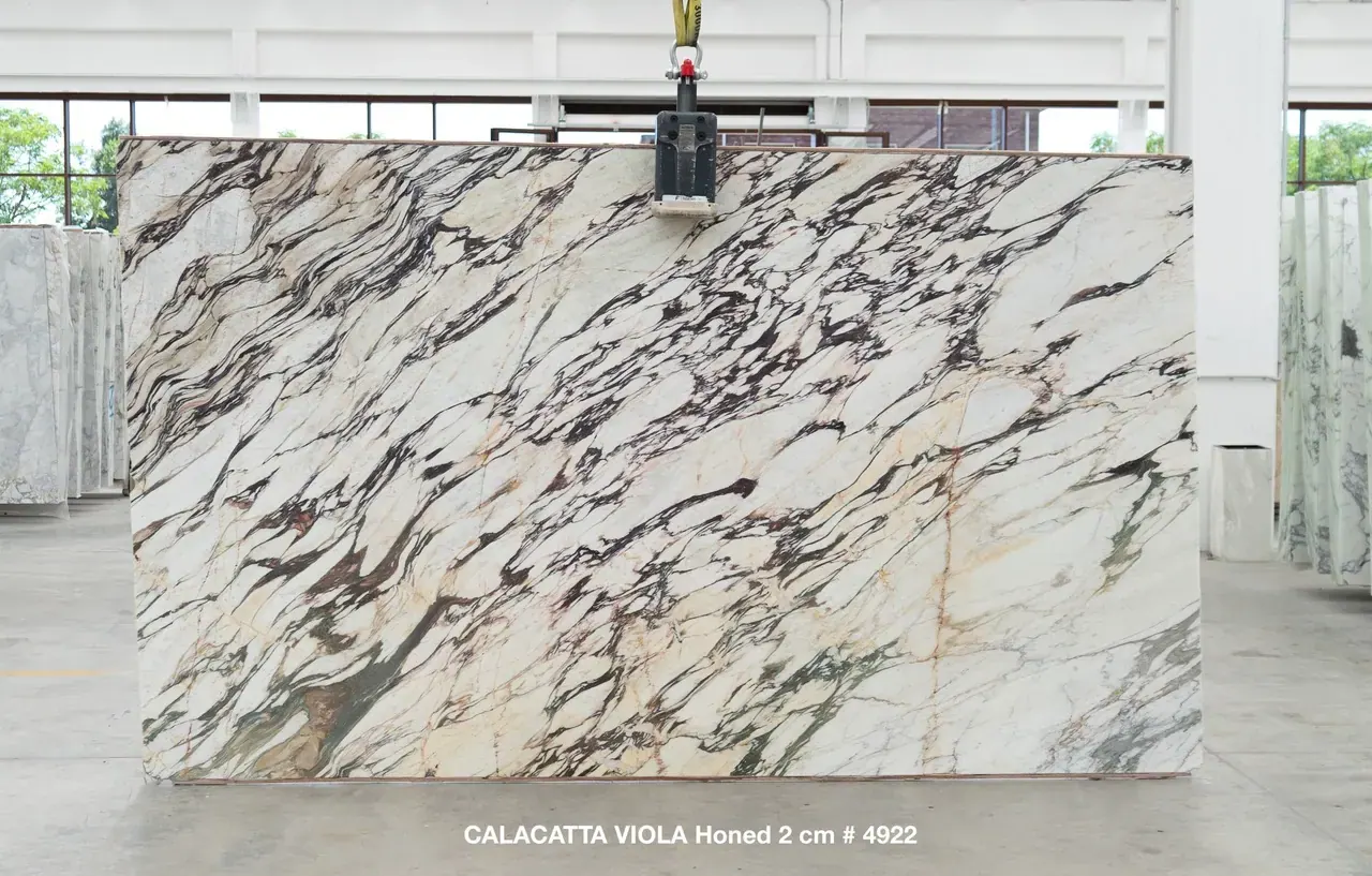 Calacatta Viola Honed 2cm marble slab, 130" × 71", lifted on overhead crane at the factory — white field crossed by diagonal violet, oxblood, and faint gold breccia veining