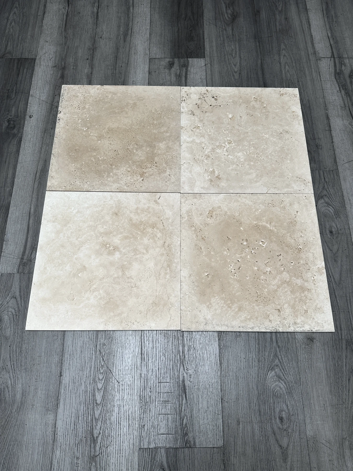 Four 18 by 18 unfilled, cross-cut and honed Roman travertine tiles laid on grey wood, showing the cream-ivory face and natural pits of in-stock yard material.