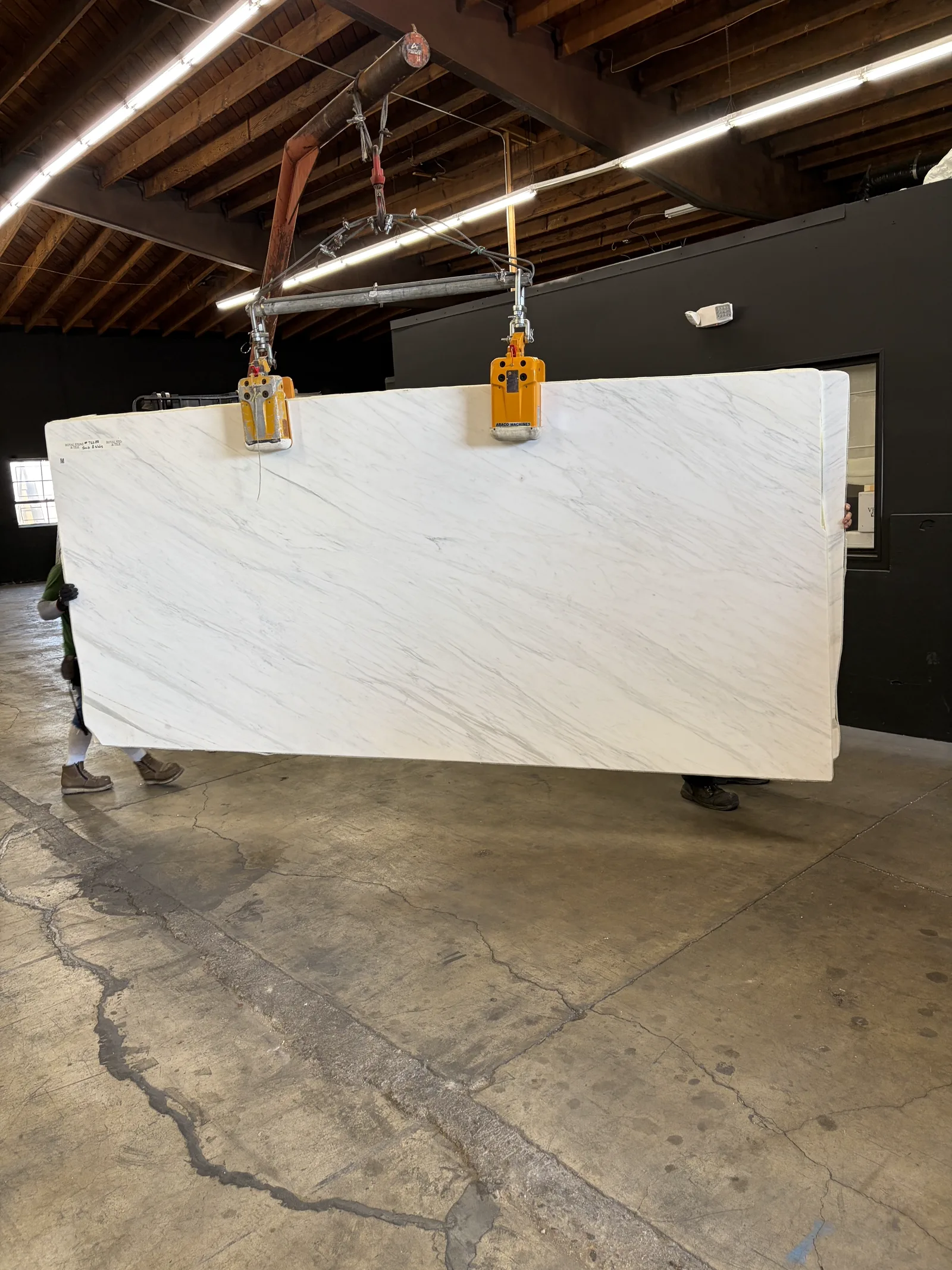 Second view of a Calacatta Caldia Soft White marble slab from lot PLST1418 carried across the Royal Stone yard floor by crane clamps, Los Angeles