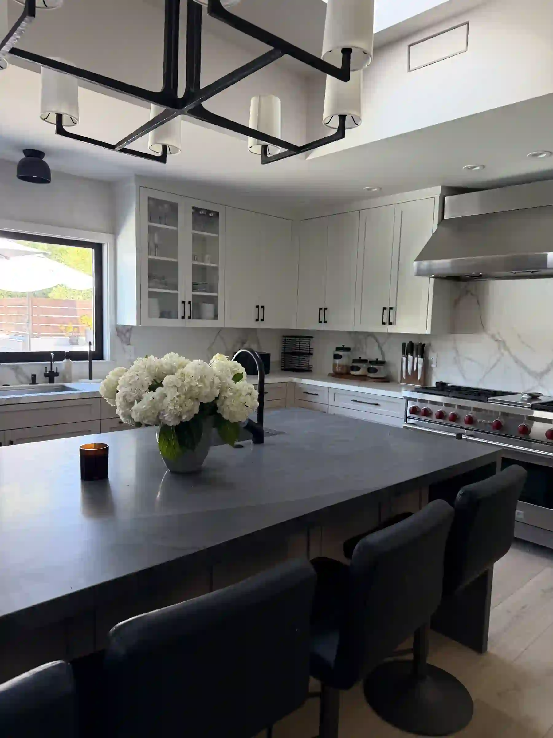 Kitchen with a large honed Black Fusion quartzite island in a deep charcoal field, paired with polished Statuary Vein Italian marble perimeter countertops and a full-height marble backsplash behind a professional range, white shaker cabinetry with black pulls, black counter stools, and a black linear chandelier.