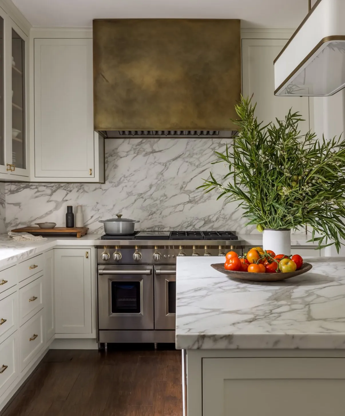 Classic white kitchen with a full-height Calacatta Borghini marble range-wall backsplash and matching island top — soft grey-and-gold veining on a bright white field — paired with pale sage shaker cabinetry, an aged-brass hood, and a stainless range.