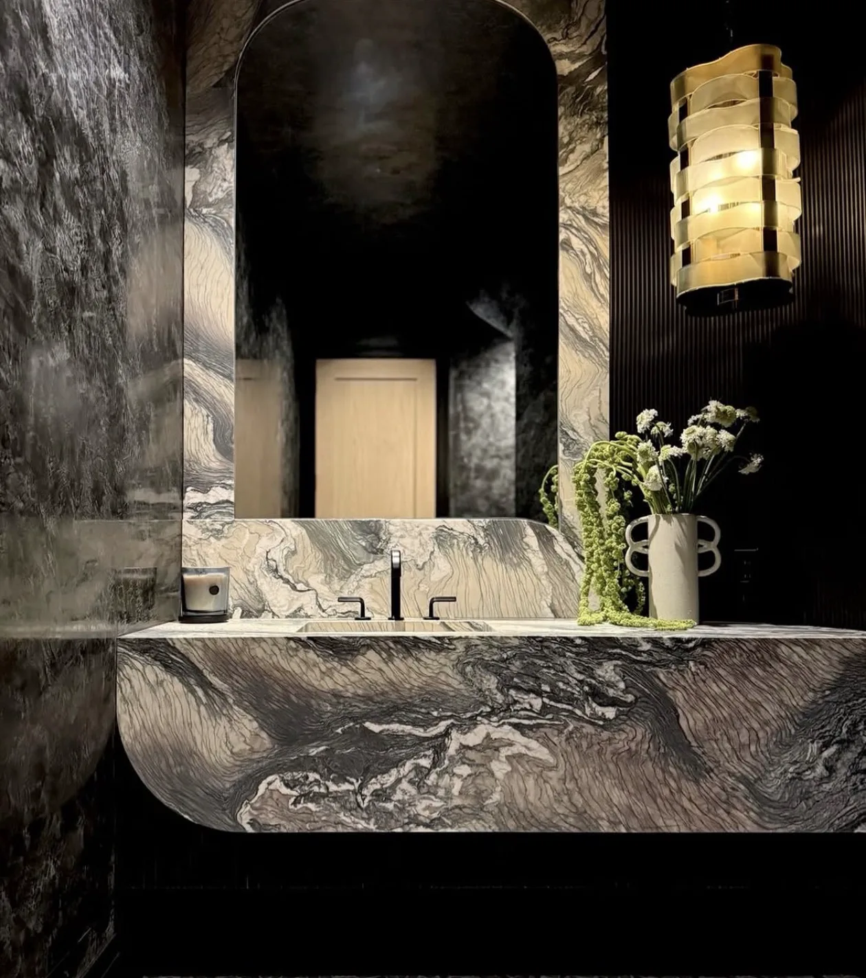 Dramatic powder room clad floor-to-ceiling in Cippolino marble — a dark grey-and-white swirled field with a monolithic stone vanity, softly radiused front, and integrated basin, a full-height arched mirror set into the bookmatched stone wall, a sculptural brass cylinder pendant, fluted dark millwork, and a ceramic vase of white blooms and trailing green amaranth.