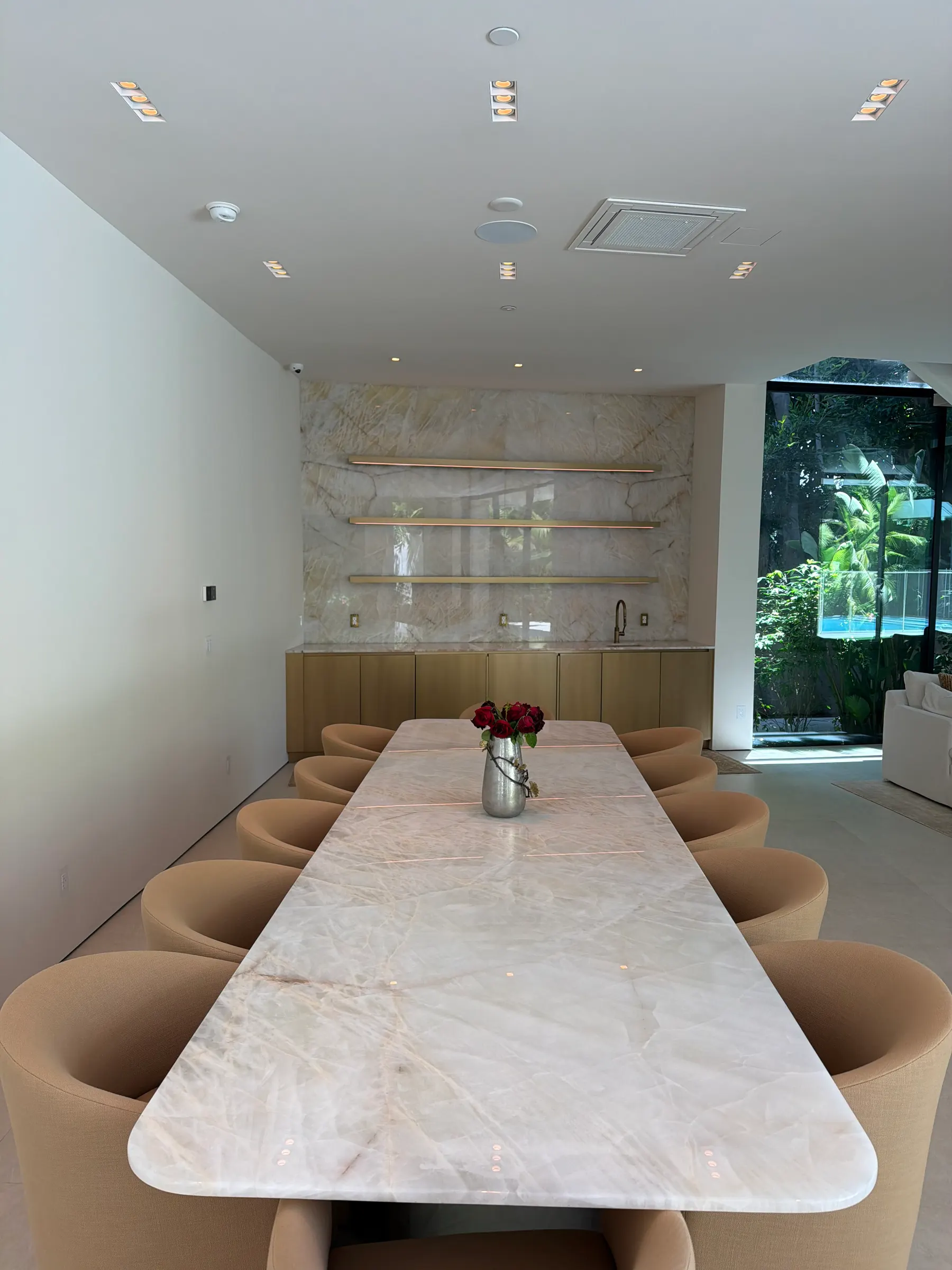 Long Cristallo quartzite dining table seating ten, set against a full-height Cristallo wet bar wall with three brass floating shelves and oak base cabinetry, opening onto a sunlit garden through floor-to-ceiling glass.