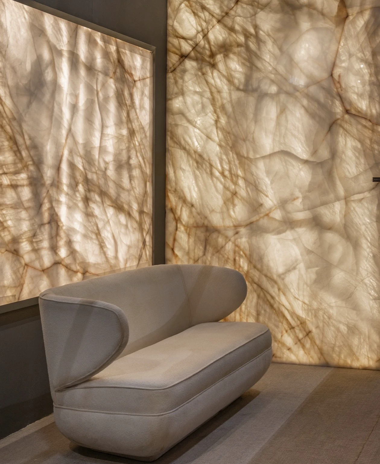 Lounge with a sculptural cream chair set before a backlit Cristallo quartzite wall by Antolini, the translucent ivory slabs crossed with warm amber veining and framed in charcoal.