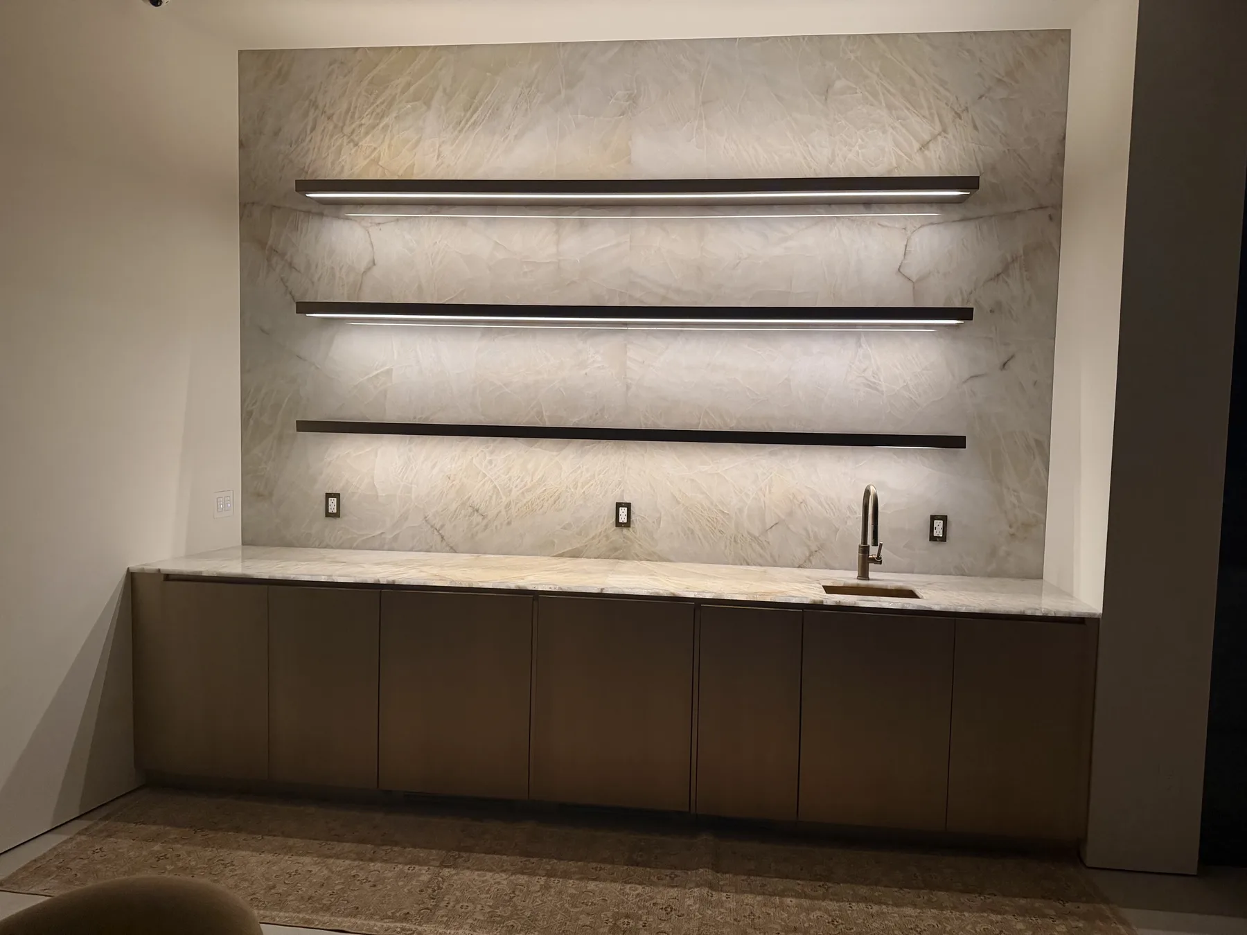 Wide elevation of a residential wet bar — full-height Cristallo quartzite back wall reading floor to ceiling, three slim bronze floating shelves with concealed linear under-lighting that backlights the crystalline field, honed Cristallo counter with a single undermount bar sink, taupe minimalist cabinetry below, and a faded antique runner on the floor.
