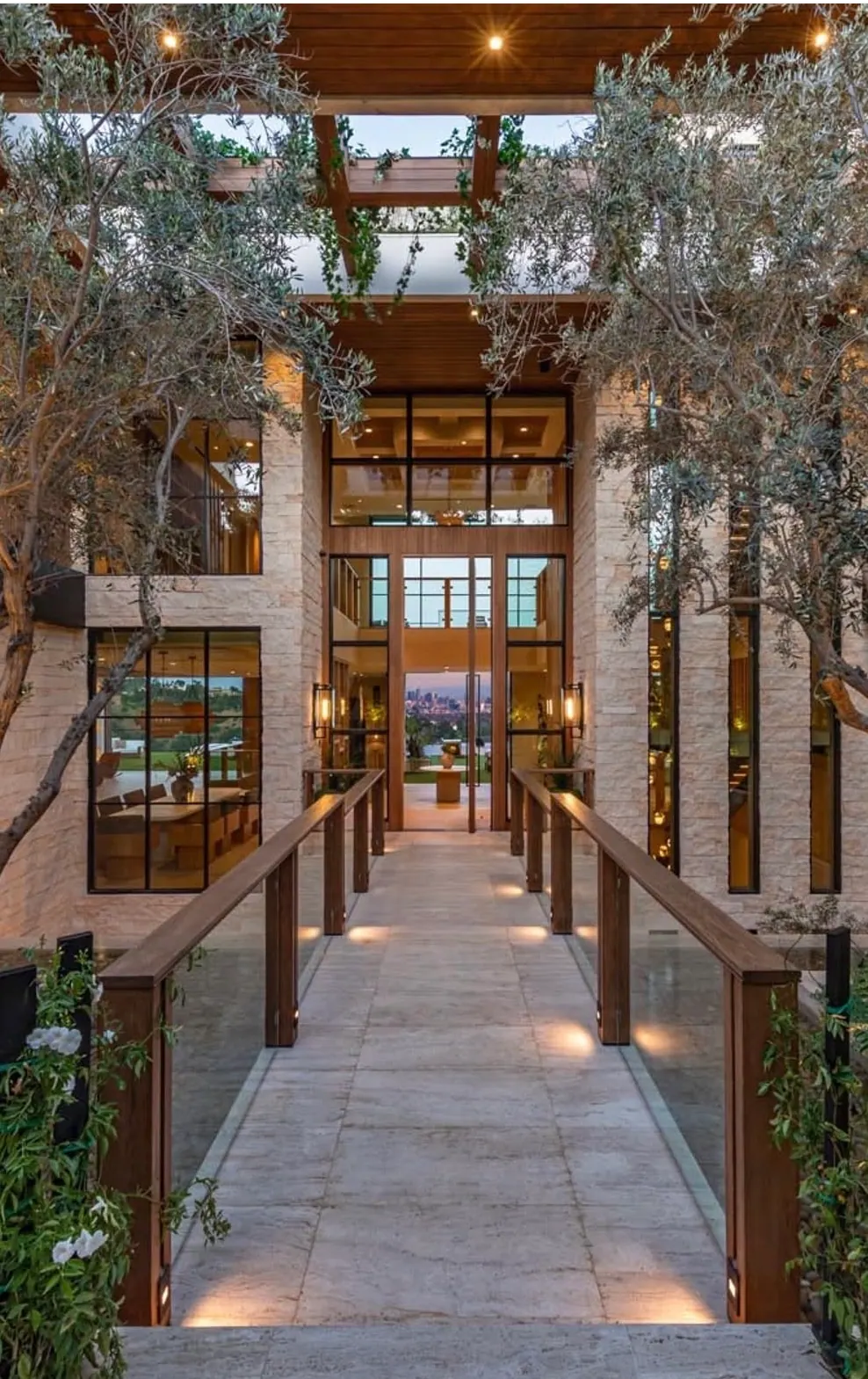Hillside residence entry bridge in vein-cut travertine pavers, flanked by olive trees and a limestone-clad steel-and-glass facade, overlooking the Los Angeles skyline at dusk.