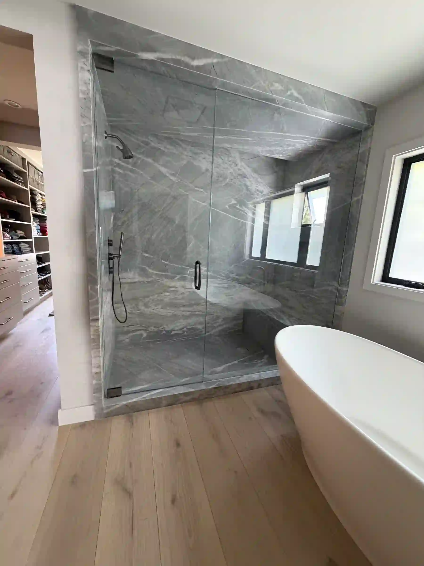 Primary bath with a frameless glass walk-in shower lined floor-to-ceiling in Antolini Gabana Nuage Bianco quartzite — a honed gray field threaded with white veining wrapping the walls, ceiling, bench, and floor, with matte-black fittings, a recessed window, a freestanding white tub, and pale oak floors.