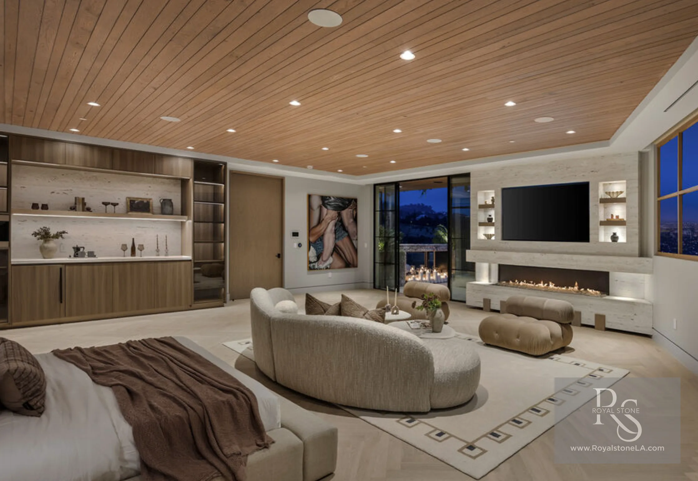 Primary suite at 1360 Laurel Way in Beverly Hills at dusk — a long horizontal cemento sandblasted travertine media wall framing a wall-mounted television above a linear gas fire, flanked by lit niche shelves, with a slatted wood ceiling, a curved bouclé sofa, and steel-framed glass doors opening to a candlelit terrace over the city lights.