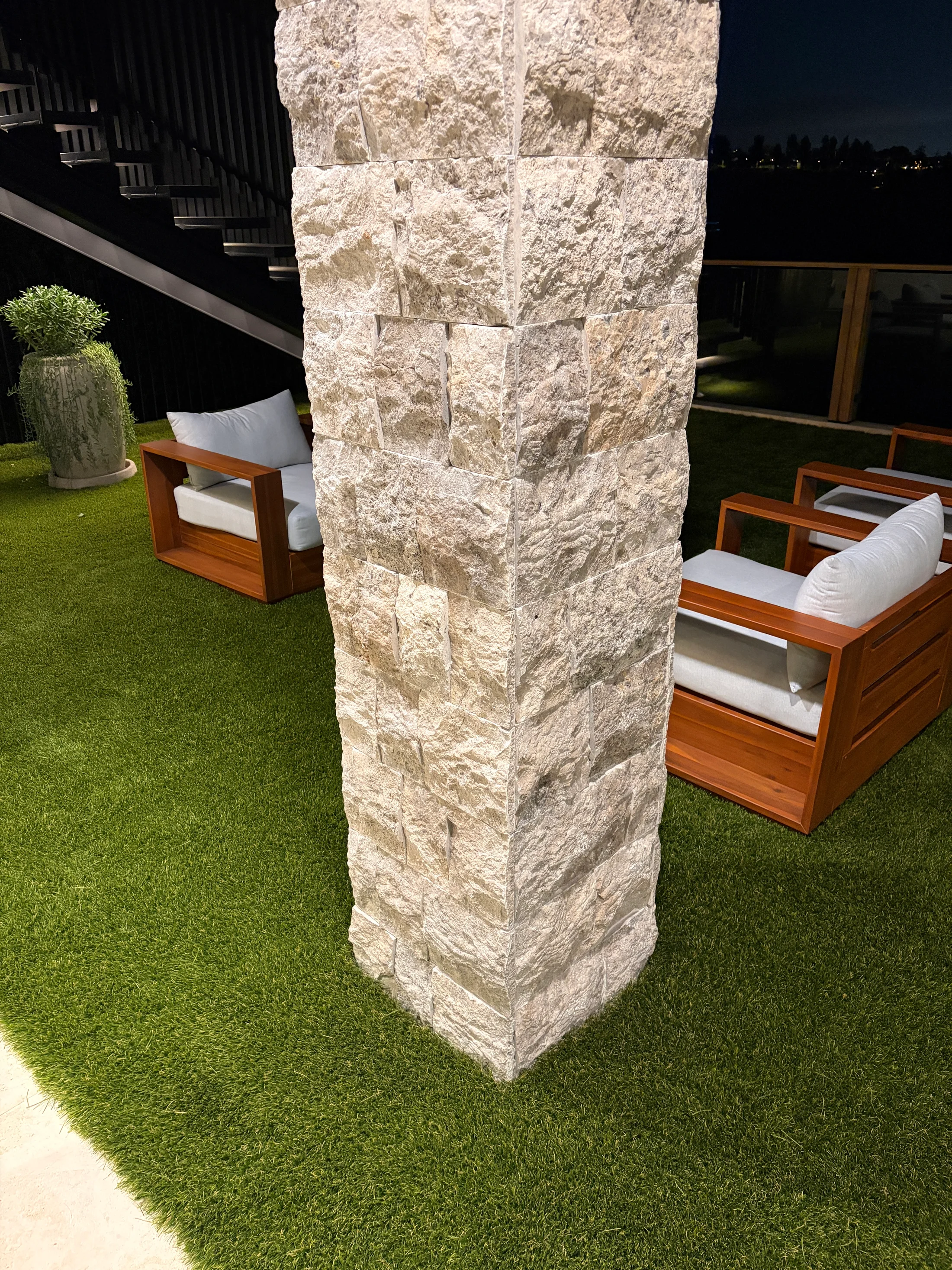 Outdoor fireplace column at 1360 Laurel Way in Beverly Hills — a freestanding pier clad on all faces in cleft-face silver travertine, set on a lawn beside teak lounge chairs beside the pool, lit at dusk with the home and Hollywood Hills behind.