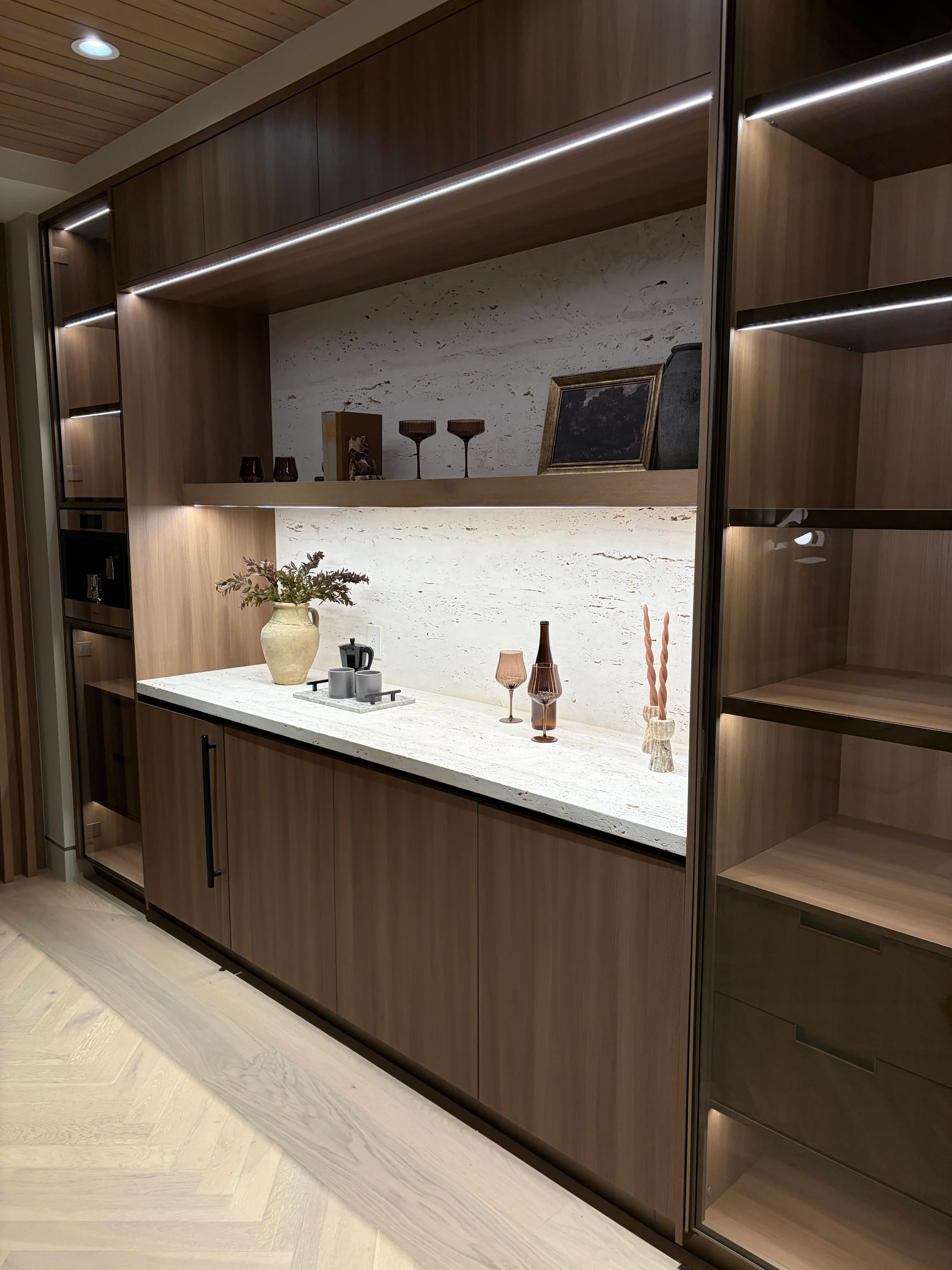 Service bar at 1360 Laurel Way in Beverly Hills — a Patagonia Tiffany quartzite countertop in a soft aqua-green crystalline field with grey-gold veining, paired with a vein-cut travertine backsplash, walnut millwork, open shelves with bottles and books lit on dimmers, and a brass below-counter basin and bridge faucet.
