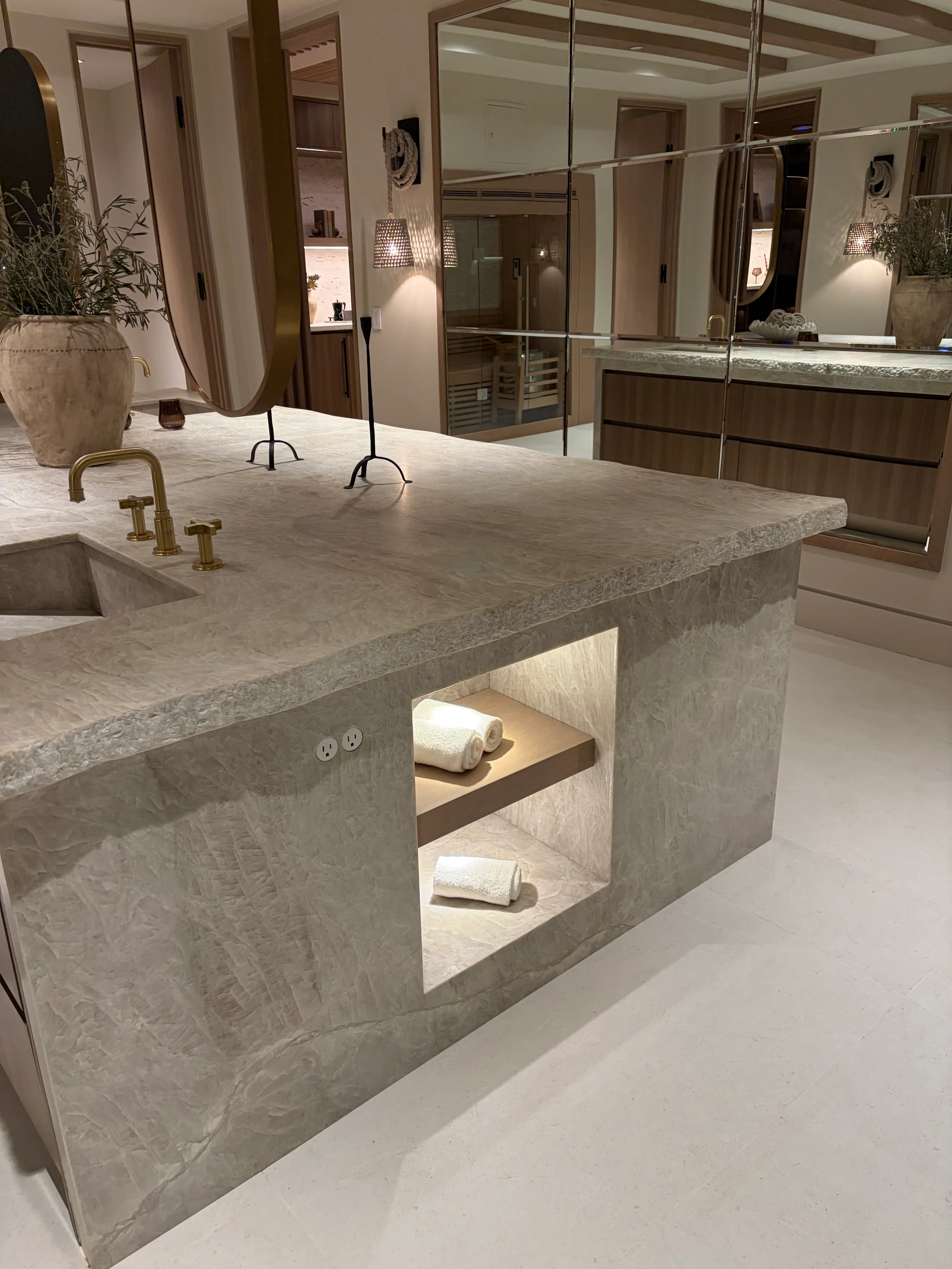 Kitchen detail at 1360 Laurel Way in Beverly Hills — the working end of a silver travertine island, a thick mitred-edge top with a recessed display niche holding decanters and ceramics, paired with an aged-brass bridge faucet, pale oak cabinetry with bronze pulls, and the open tread of a sculptural wood stair in the background.