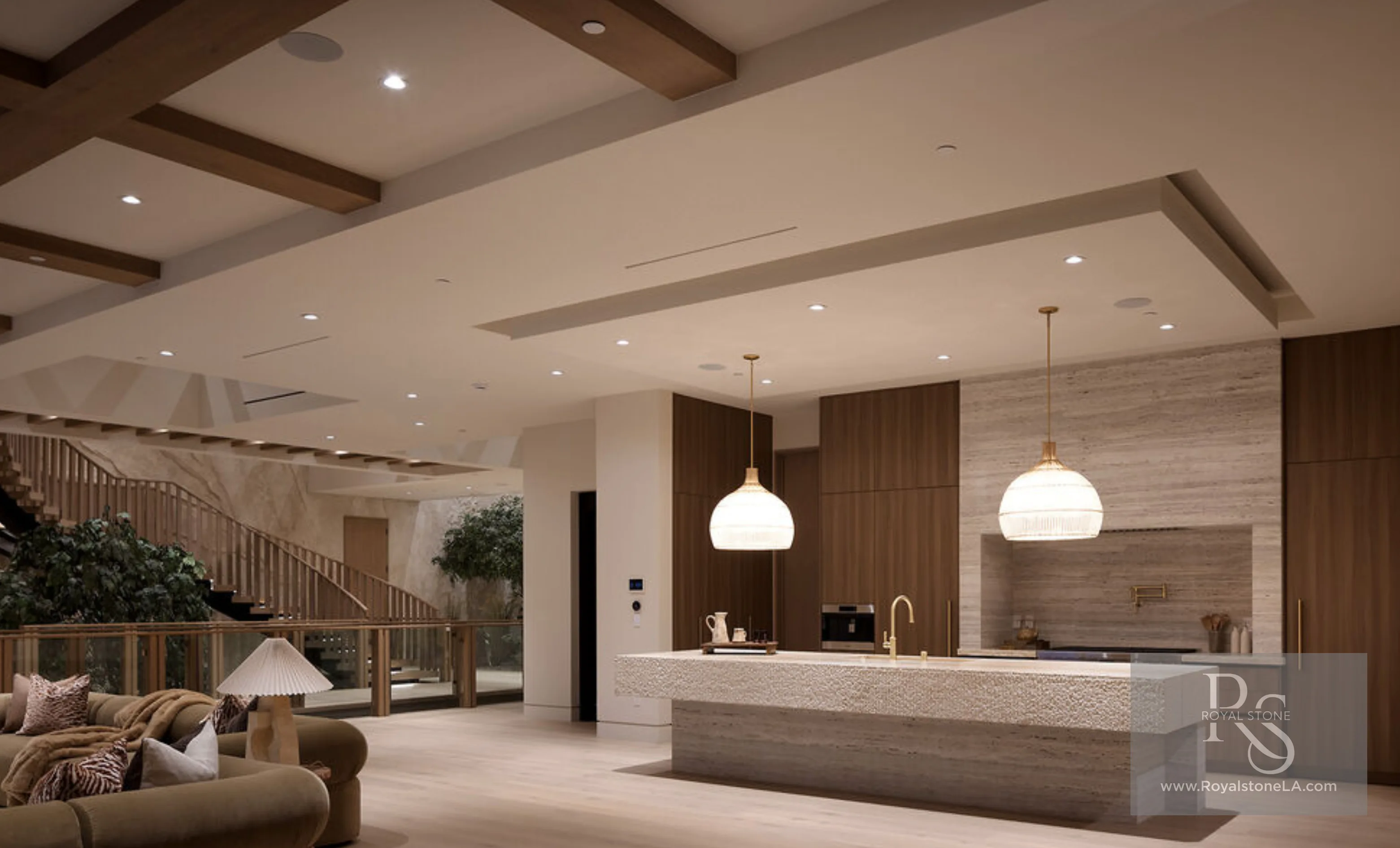 Evening kitchen at 1360 Laurel Way in Beverly Hills — a long silver travertine island with a hand-chiseled, combed apron and matching full-height range-wall backsplash in a soft vein-cut field, paired with walnut slab cabinetry, woven globe pendants, an open wood stair, and pale oak floors.