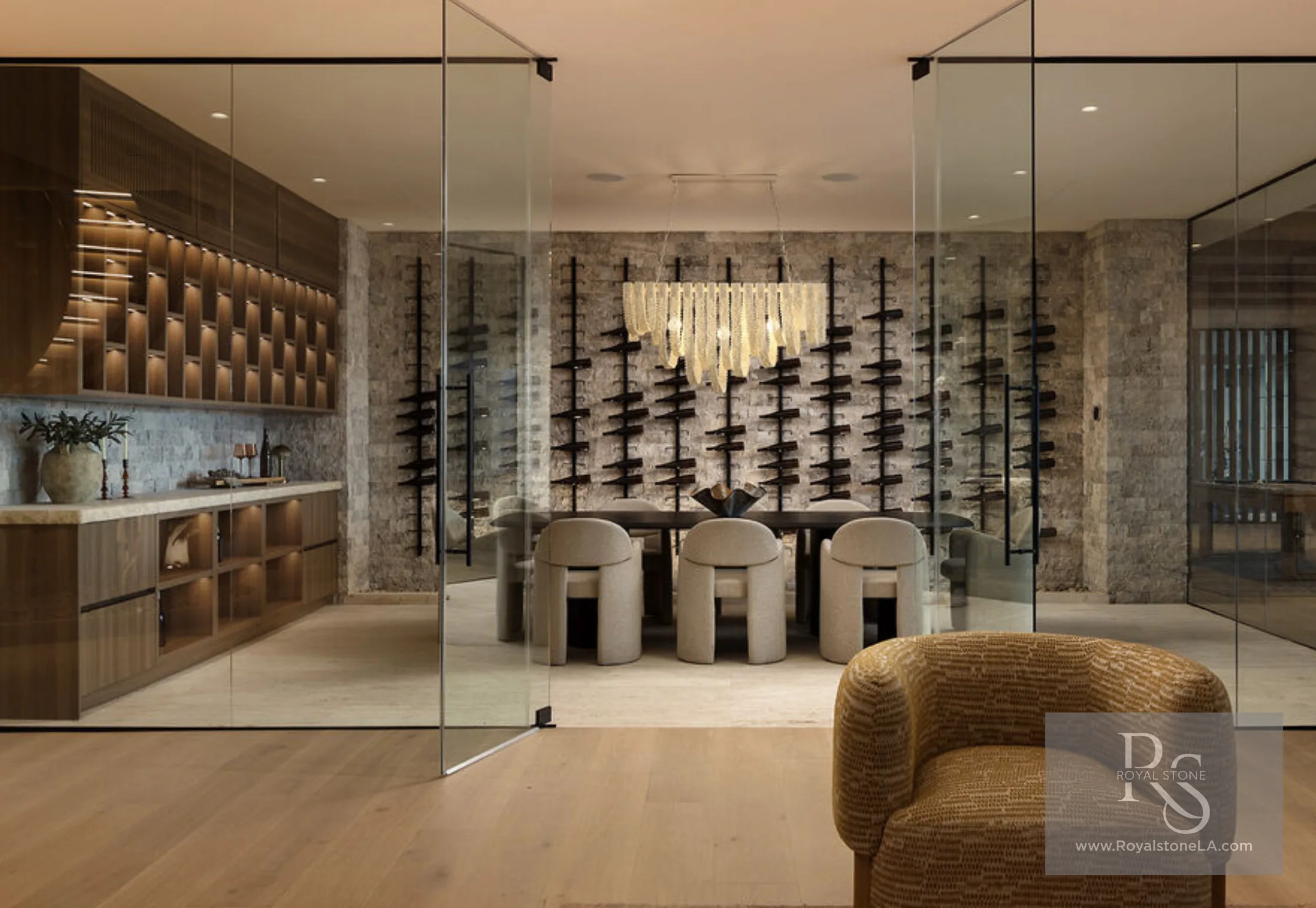Glass-walled wine and dining room at 1360 Laurel Way in Beverly Hills — full-height split-face cleft travertine walls behind black steel bottle racks, a long dining table with sculptural bouclé chairs under a cascading glass chandelier, viewed through frameless glass partitions from an adjoining walnut bar.