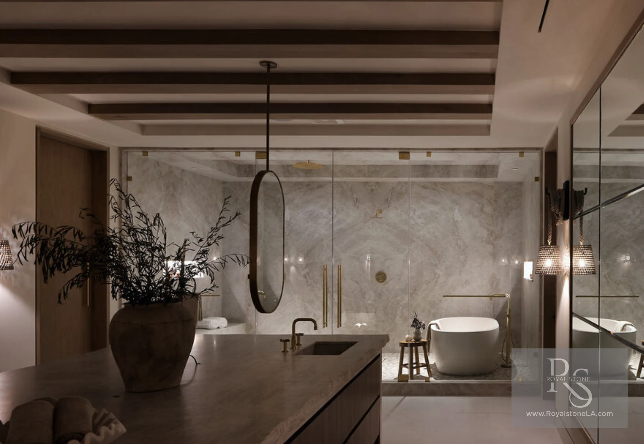 Primary bath at 1360 Laurel Way in Beverly Hills at night — a glass-enclosed wet room lined floor-to-ceiling in bookmatched Taj Mahal quartzite with soft ivory veining, a freestanding tub with an aged-brass floor filler, a brass rain head, and a stone-topped vanity island with a sculptural urn, under wood-beamed ceilings.