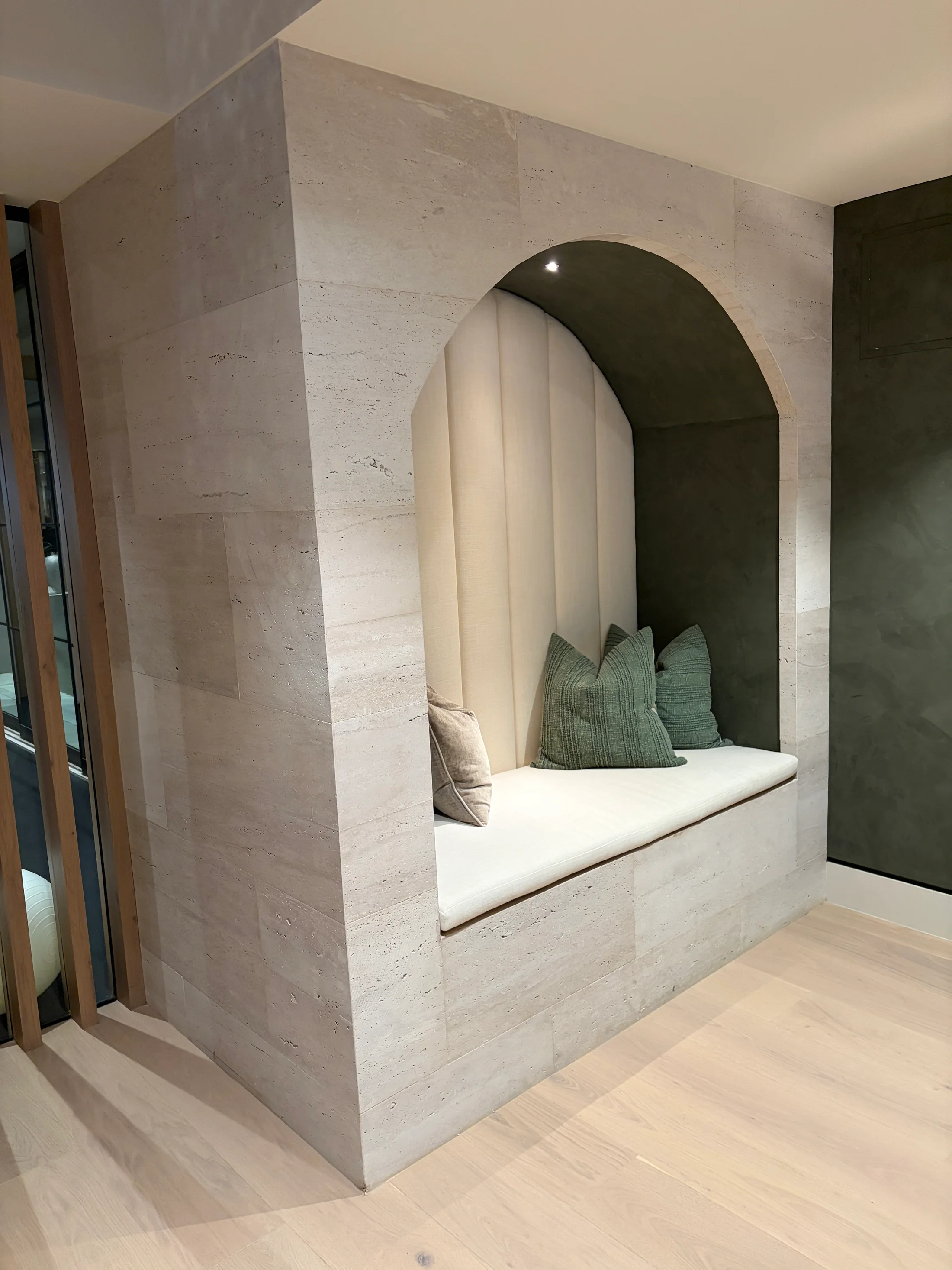 Quiet sit-down at 1360 Laurel Way in Beverly Hills — a monolithic travertine banquette carved from a single cream block, with a curved upholstered seat in ivory bouclé, olive linen cushions, and a reading light set into the stone, overlooking pale oak chevron floors and a stacked stone wall beyond.