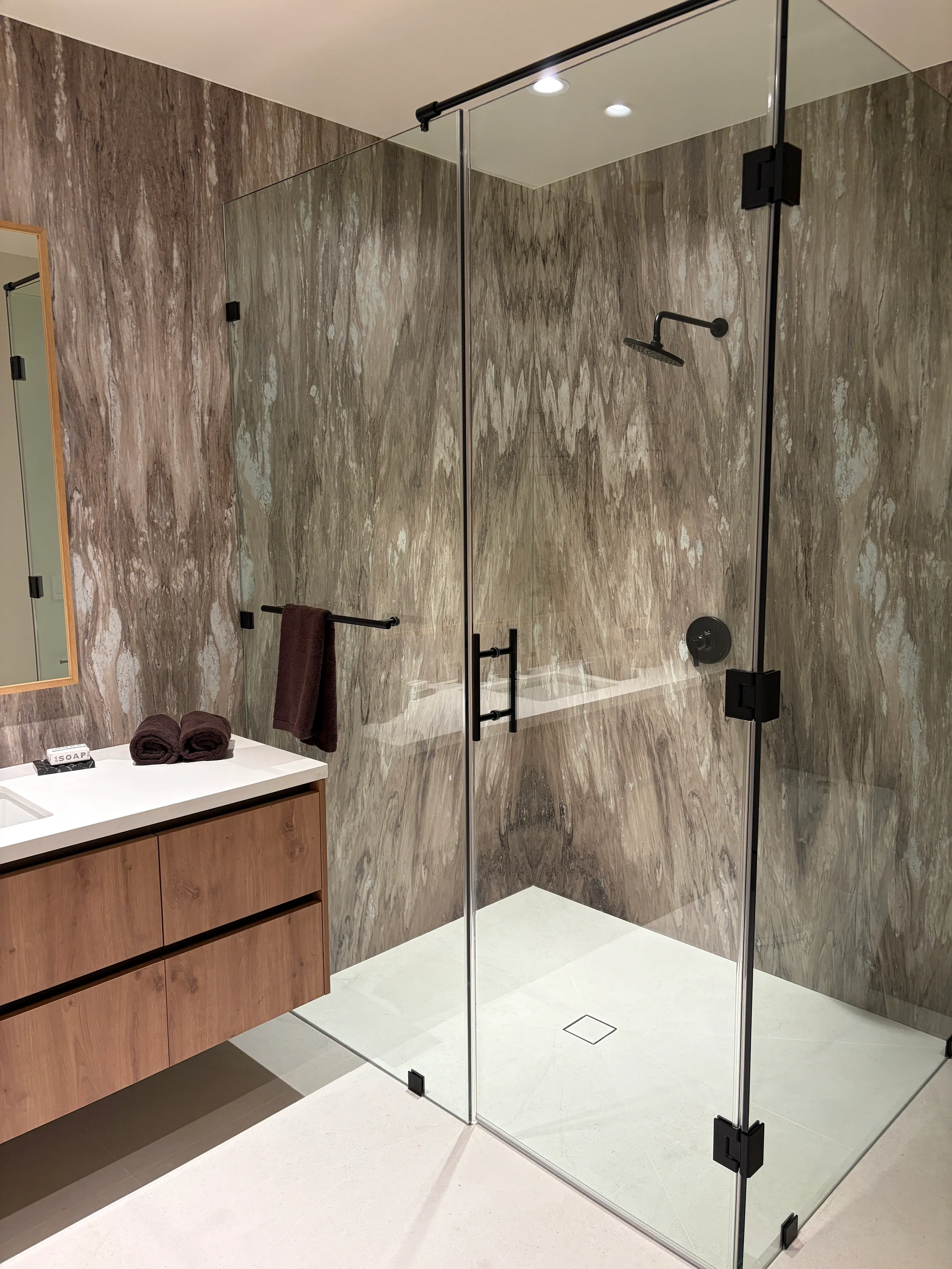 Primary shower at 1360 Laurel Way in Beverly Hills — a glass-enclosed wet room lined floor-to-ceiling in vein-cut travertine in a cool grey-and-ivory banded field, a teak bench seated beneath a long ceiling-mounted rain head, matte-black fittings, and a frameless glass partition opening onto the suite.