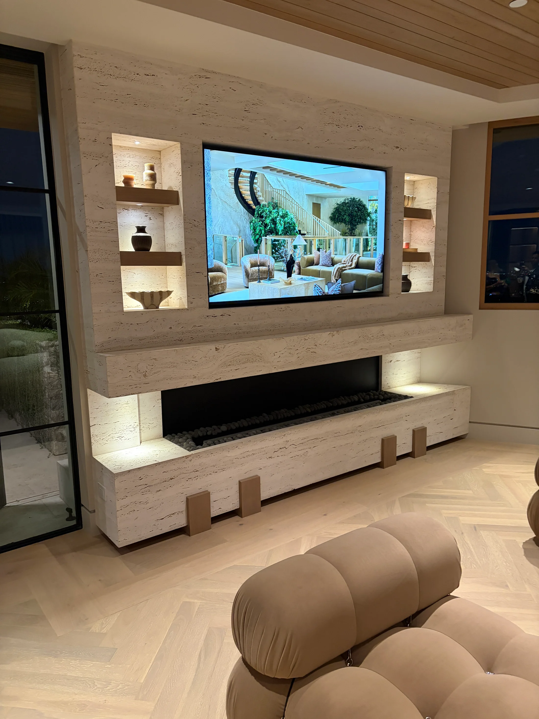 Lounge at 1360 Laurel Way in Beverly Hills — a vein-cut travertine media wall framing a recessed television above a long linear fire, flanked by an inset niche grid holding books and ceramics, with a curved bouclé lounge chair and slatted wood ceiling above pale oak chevron floors.