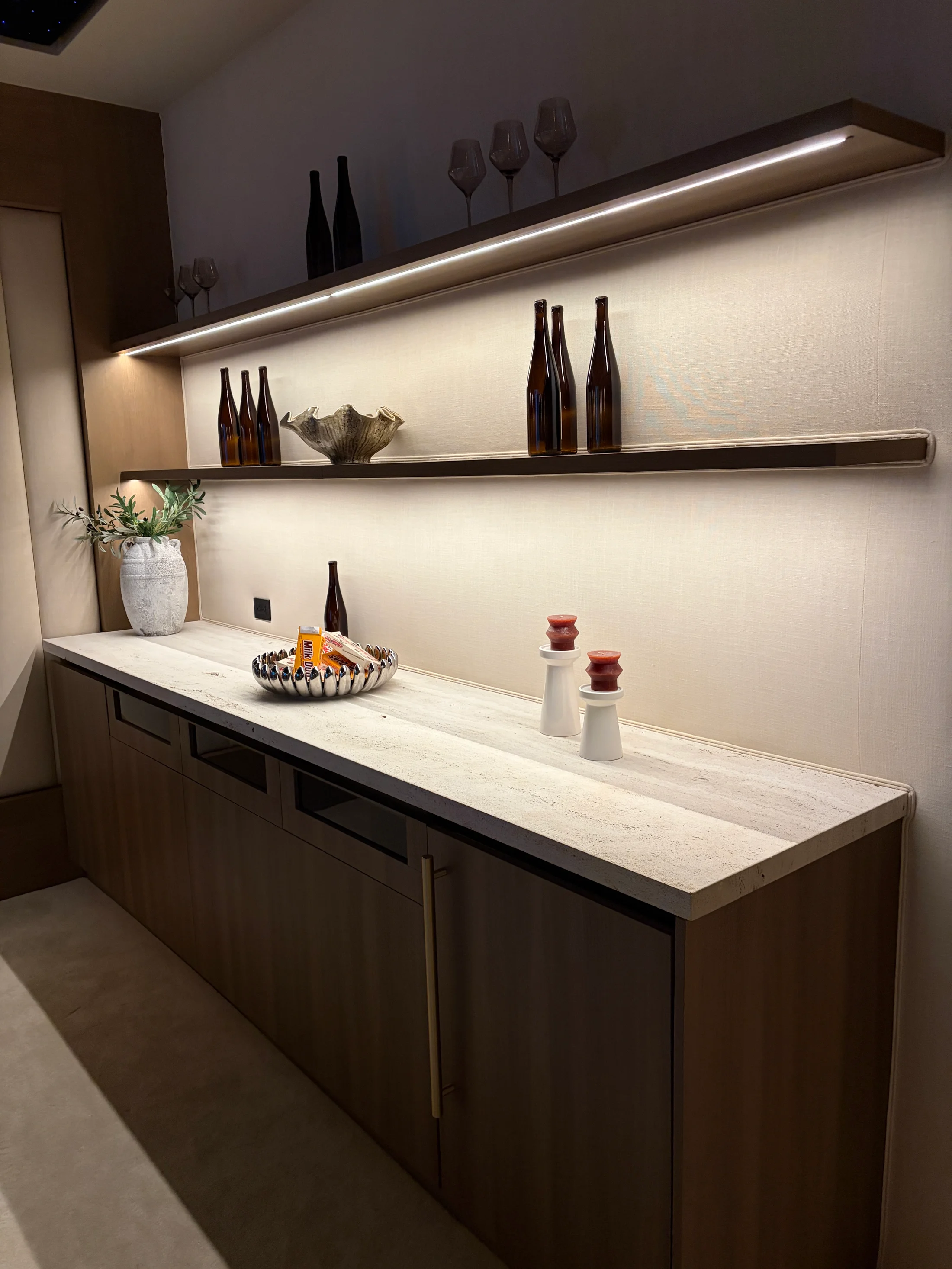 Powder room bar at 1360 Laurel Way in Beverly Hills — a vein-cut travertine back wall and counter paired with floor-to-ceiling walnut millwork, brass pulls, a recessed display niche lit on dimmers holding bottles and stacked ceramics, and a tone-on-tone bouclé stool pulled up to the under-mounted basin.