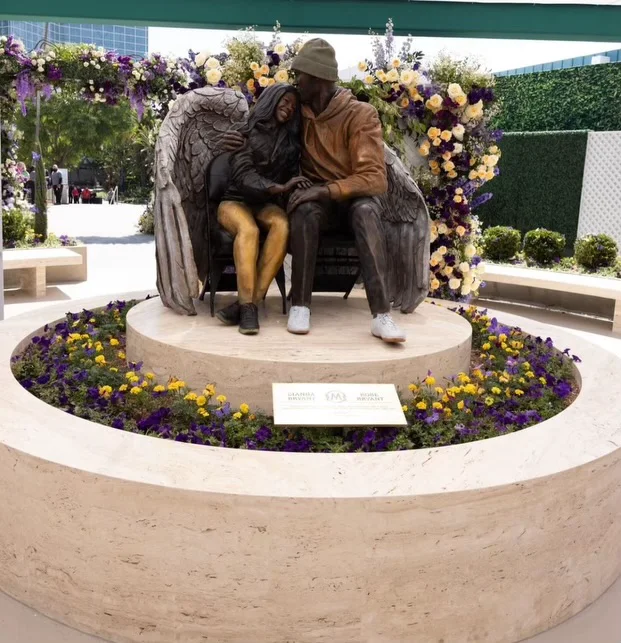 Outdoor memorial — a bronze statue of Kobe and Gianna Bryant seated together, mounted on a circular Navona travertine plinth and surrounding bench in a soft ivory unfilled field, ringed by purple and gold flowers.