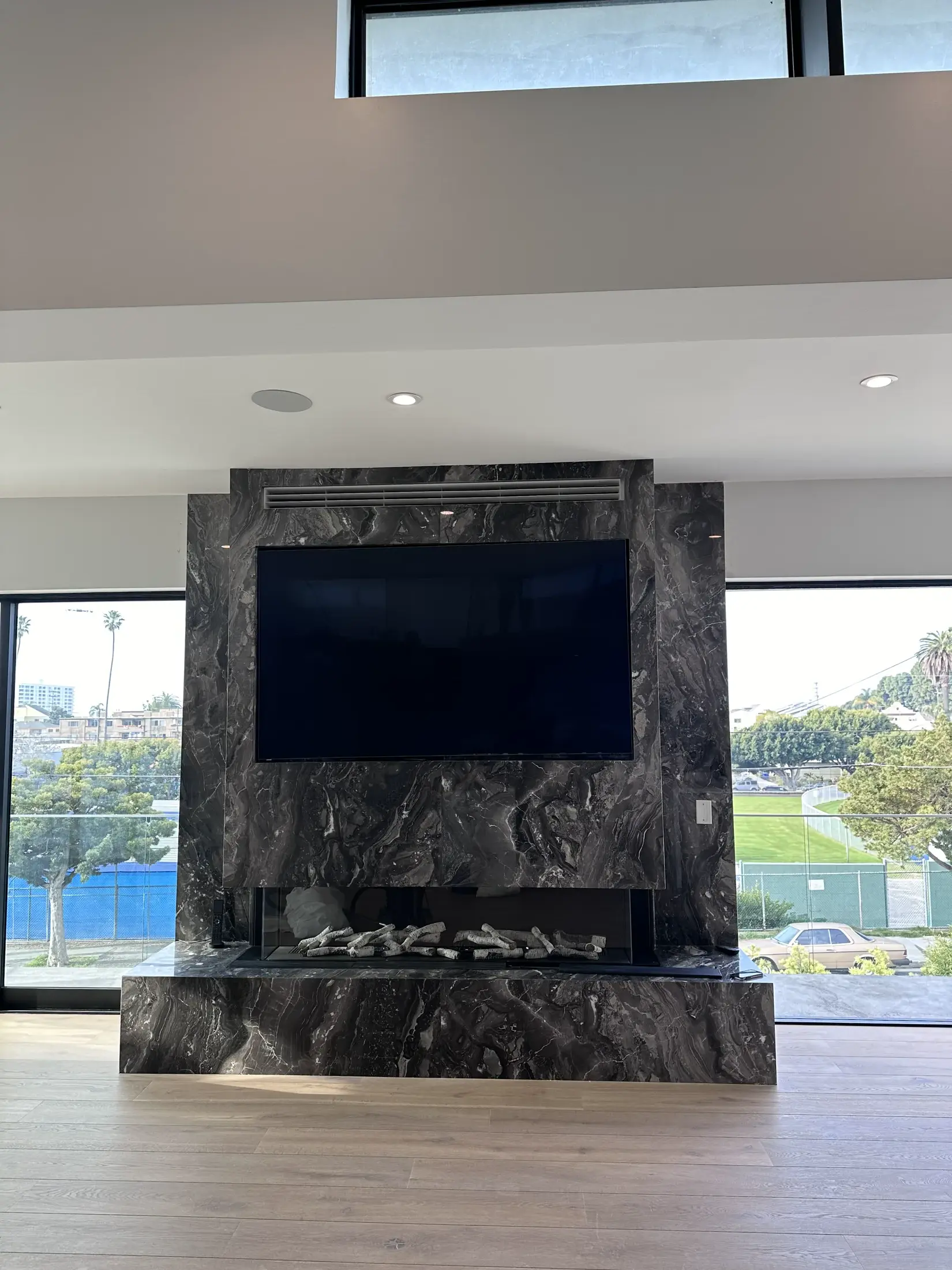 Italian Nero Orobico marble fireplace at a Los Angeles residence — a dramatic black-and-white flowing-veined field running floor-to-ceiling across the fireplace wall, a wall-mounted television inset above a recessed linear hearth filled with stacked river stones and a sculptural branch, set against white plaster walls, recessed ceiling lights and a clerestory above, oak floors, and steel-framed glass onto a garden with palms and a tennis court beyond.