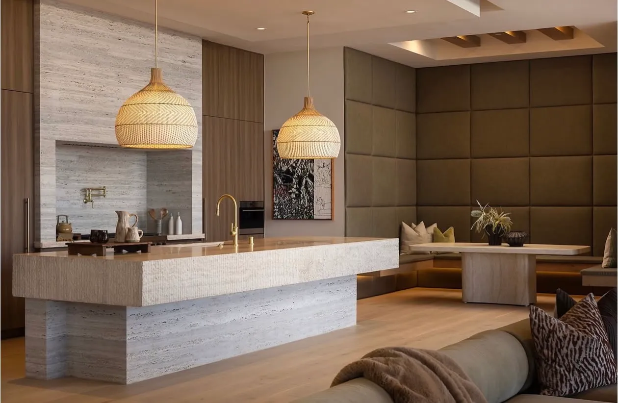 Kitchen at The Opal in Beverly Hills — a monolithic travertine waterfall island with a chiseled, combed face and matching full-height backsplash in a soft silver-ivory vein-cut field, paired with walnut slab cabinetry, an upholstered olive banquette, woven rattan pendants, and pale oak floors.