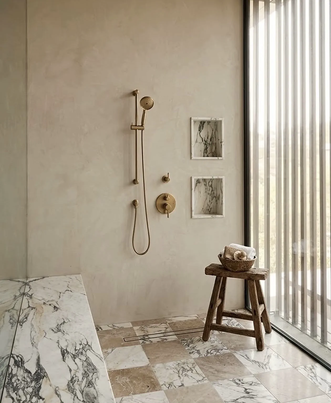 Primary bath by Ryan Saghian at 10850 Portofino Pl in Los Angeles — a warm plastered shower room with brushed-brass fittings, a checkerboard floor of Viola marble and travertine, a marble slab leaning at the edge of the frame, and a full-height window with sheer vertical blinds.