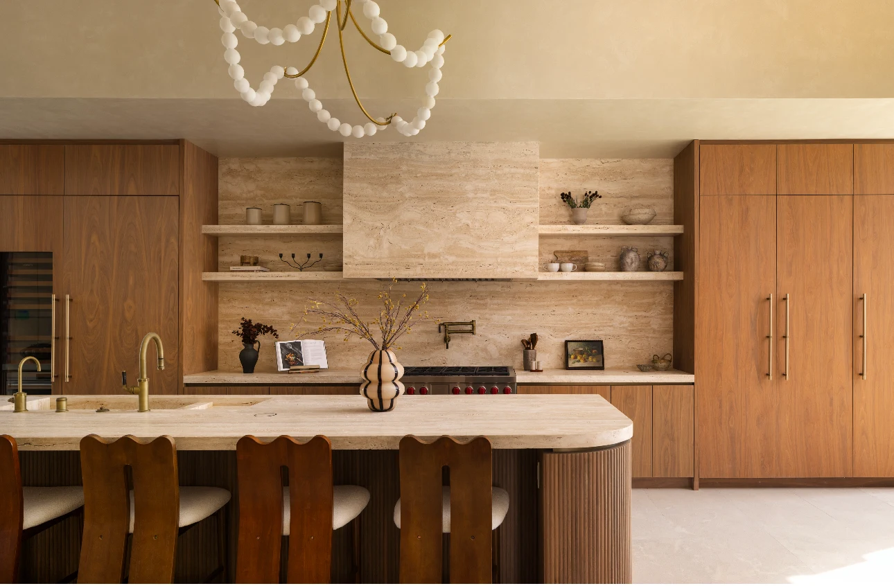 Warm kitchen by designer Ryan Saghian at Portofino in Bel-Air, created with Italian Roman unfilled travertine slabs across the island, backsplash, open shelving, and range hood, set against walnut cabinetry and sculptural wood chairs.