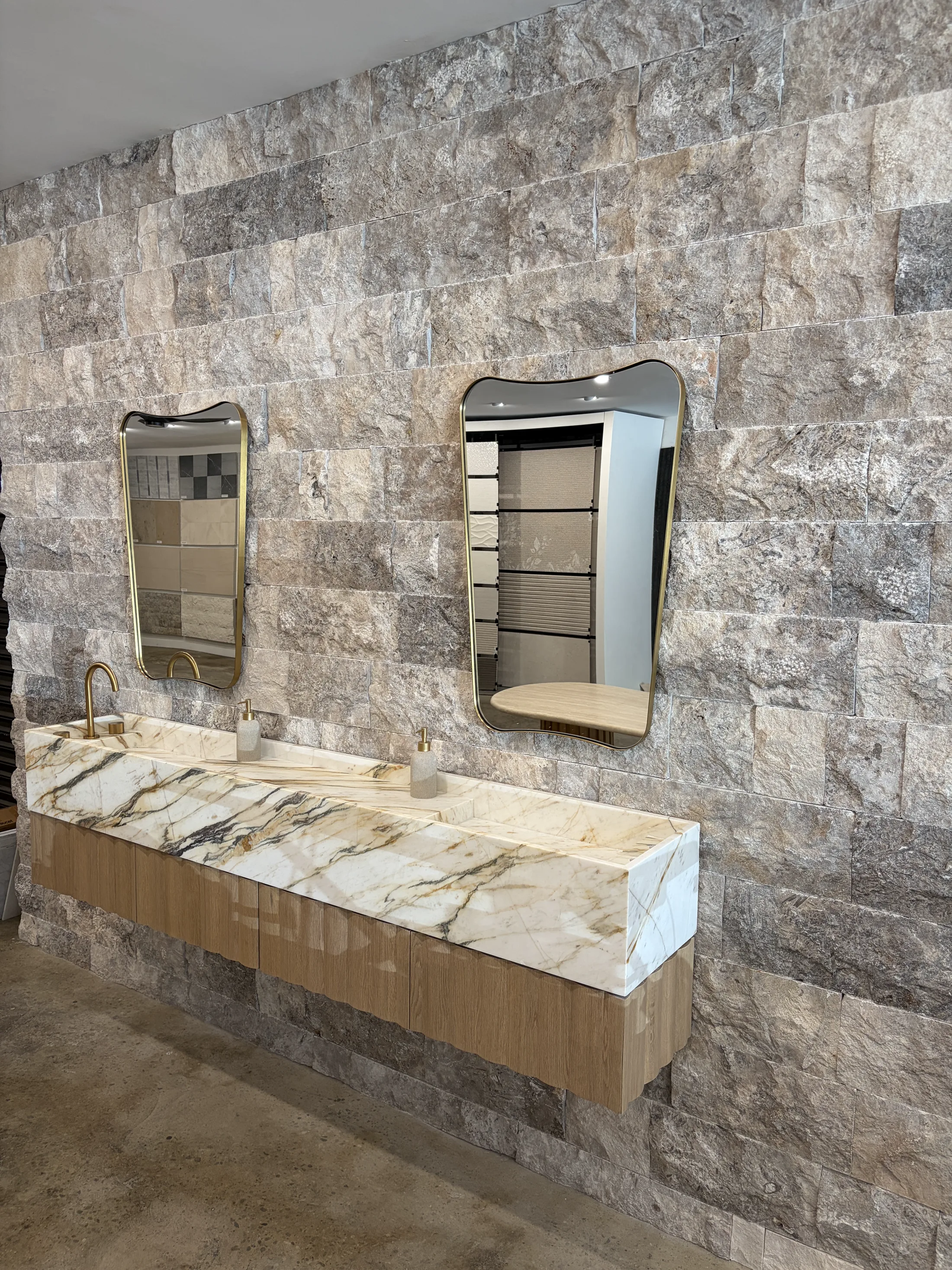 Royal Stone showroom display pairing an 8" silver travertine cleft-face backsplash — bold silver and grey fractured quarry blocks running floor-to-ceiling — with a Calacatta Monet Oro honed marble integrated trough sink and warm oak base cabinetry, brass bridge faucet, two wavy brass-framed vintage mirrors hung against the stone wall, and a polished concrete floor.