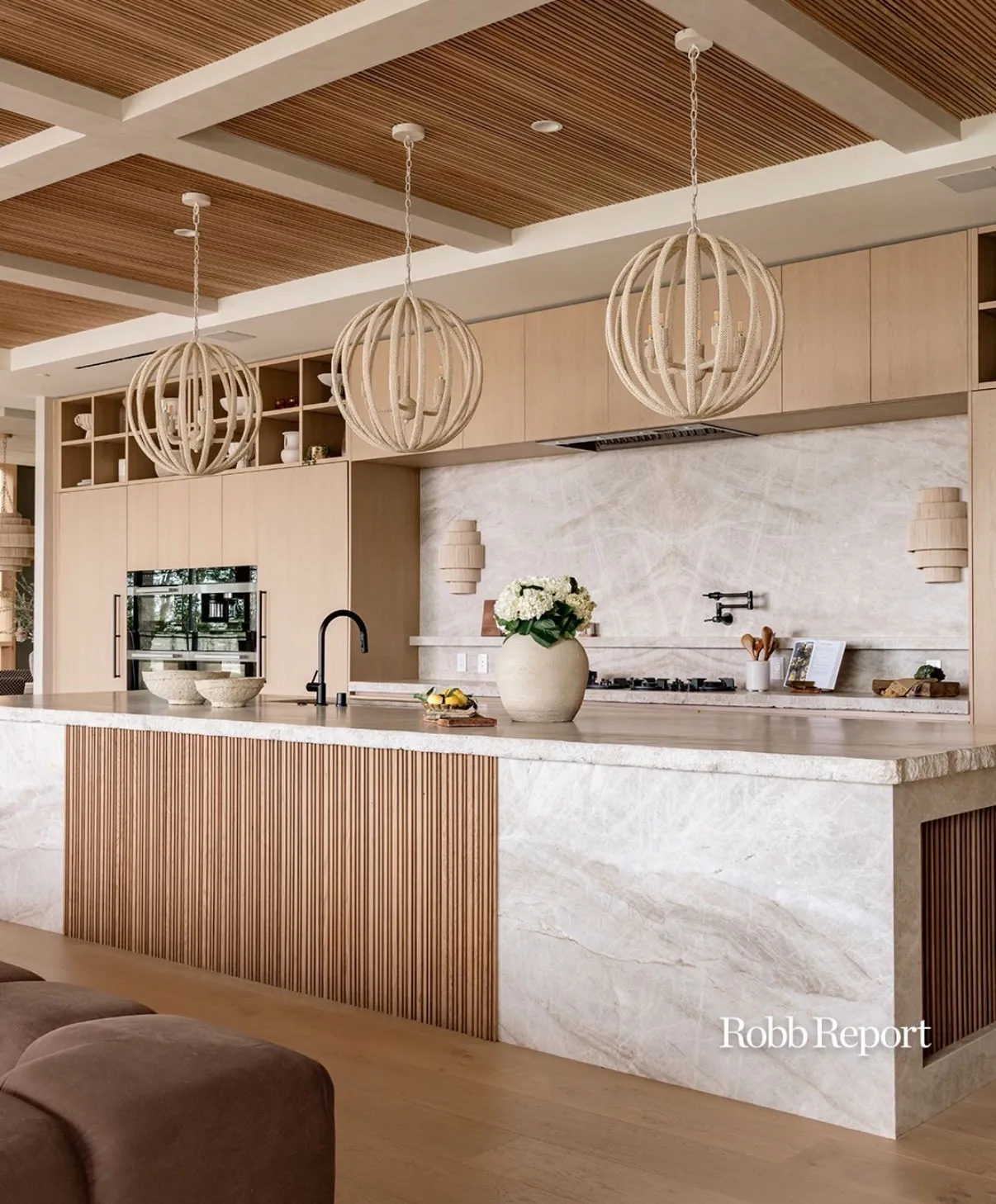 Sunlit kitchen with a long Taj Mahal quartzite island and matching full-height range-wall backsplash — soft ivory field with gentle taupe veining, a dramatic natural broken edge left raw on the island, fluted white-oak base millwork, a rope-orb pendant trio, and a slatted wood ceiling above.