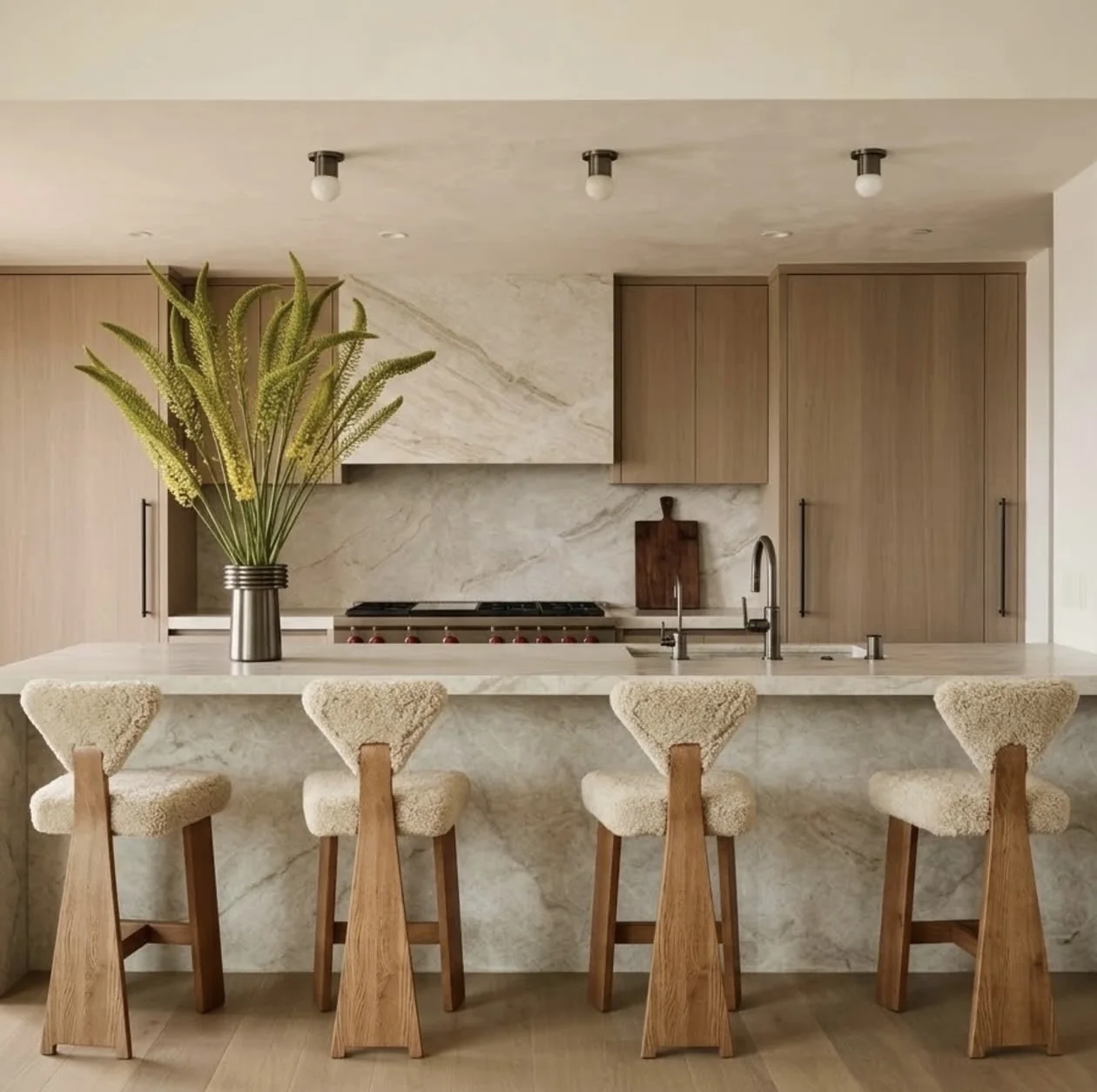 Warm contemporary kitchen by Ryan Saghian with Taj Mahal quartzite carried across the island, waterfall end, and range-wall backsplash, paired with oak cabinetry, sculptural boucle stools, and a vase of green stems.