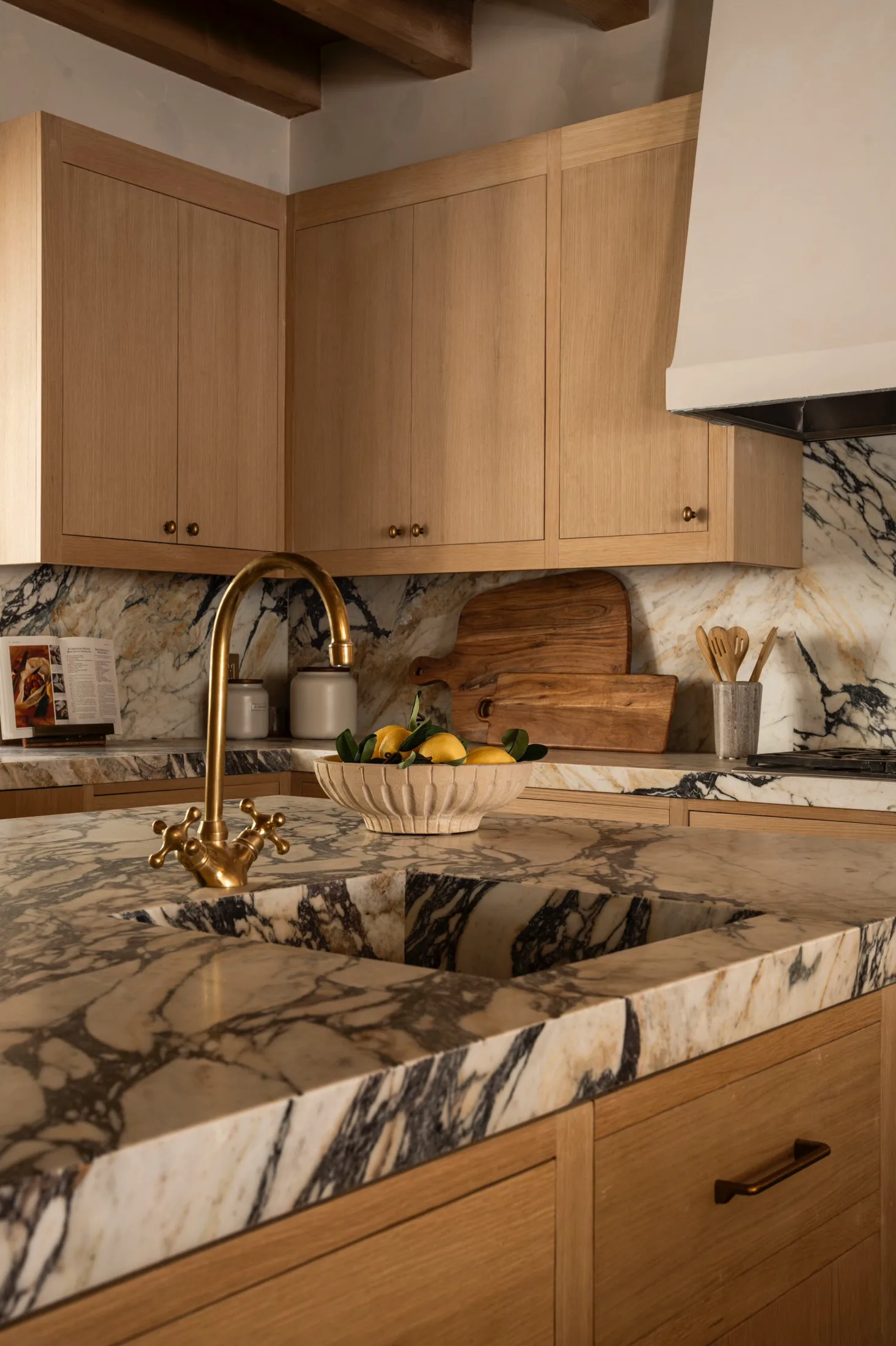 Close view of a Calacatta Viola Extra marble island — a thick mitred-edge top with a carved integral apron sink in a dramatic field of plum and charcoal veining, an aged-brass cross-handle bridge faucet, a fluted bowl of lemons, white-oak cabinetry, and a full-height marble backsplash beyond.