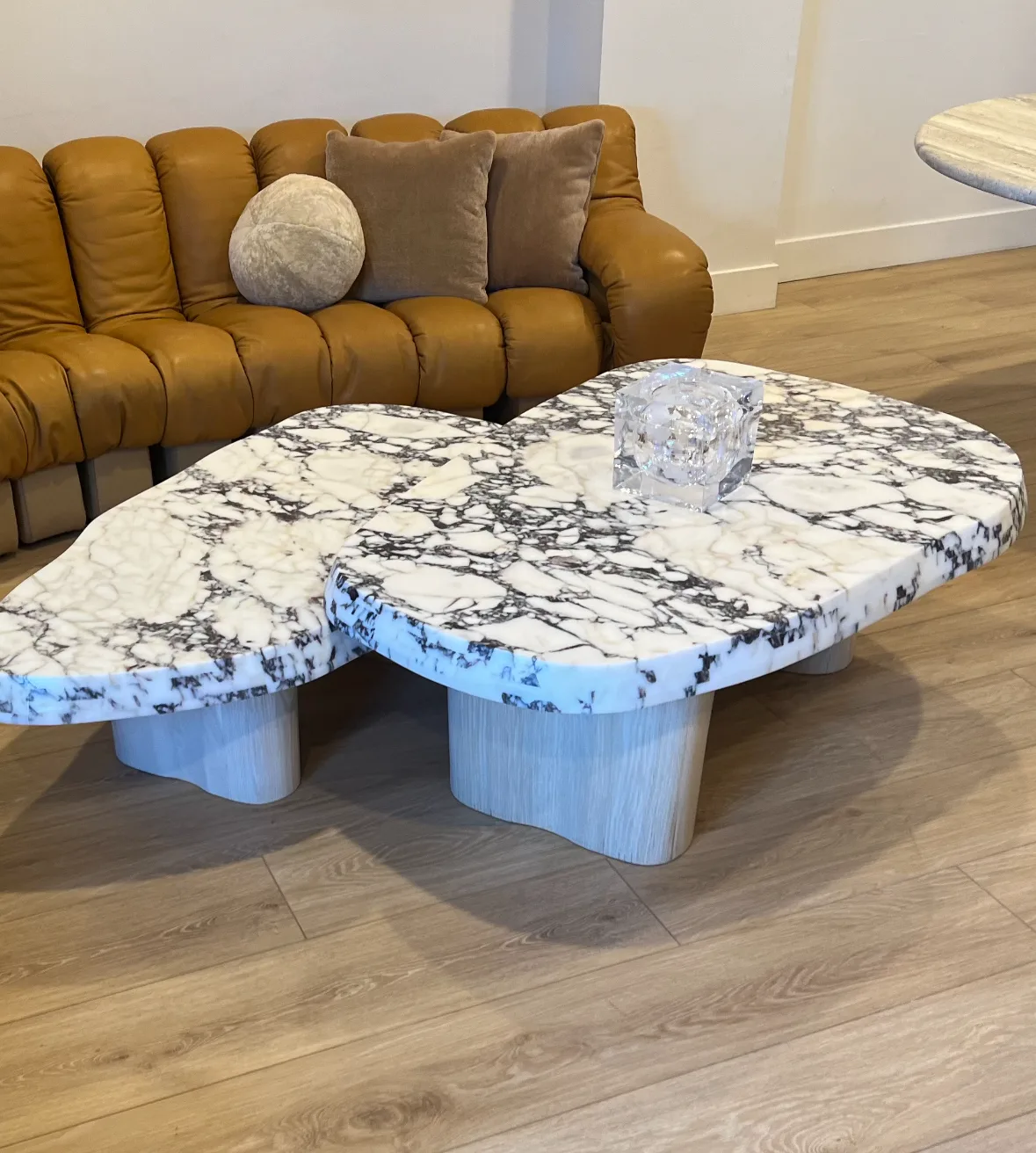 A pair of organic kidney-shaped coffee tables with honed Viola marble tops in a white-and-plum veined field, set on pale sculpted wood pedestals over oak floors, against a caramel leather tufted sectional.