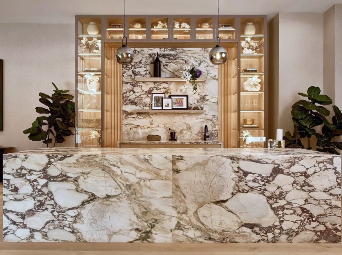 Bar at a private residence in Los Angeles — a monolithic Calacatta Viola marble waterfall island with a thick bookmatched top in a dramatic plum-and-bone veined field running across the counter, down both waterfall faces, and into the floor, paired with a matching Viola marble back wall, oak millwork cabinets with lit display niches holding ceramics and sculpture, brass globe pendants, and fiddle-leaf figs flanking the bar opening.