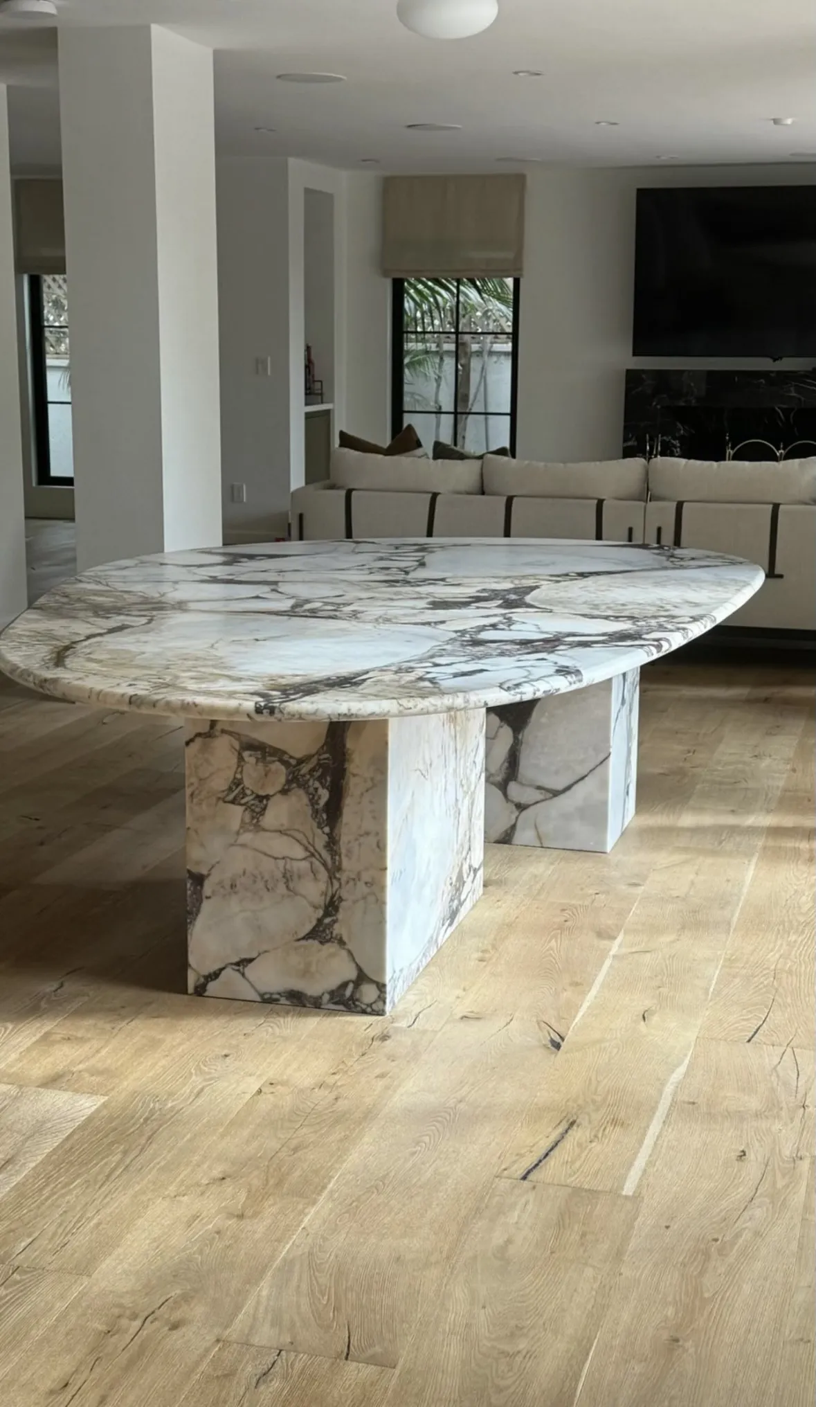 Custom Italian Calacatta Viola marble dining table in a Los Angeles residence — a soft-toned organic-form honed top with a flowing continuous edge sitting on two monolithic Viola pedestal legs, set on pale wide-plank oak floors in front of a bouclé sectional sofa under a constellation of glass globe pendants, with a walnut window valance and steel-framed glass onto the back garden.