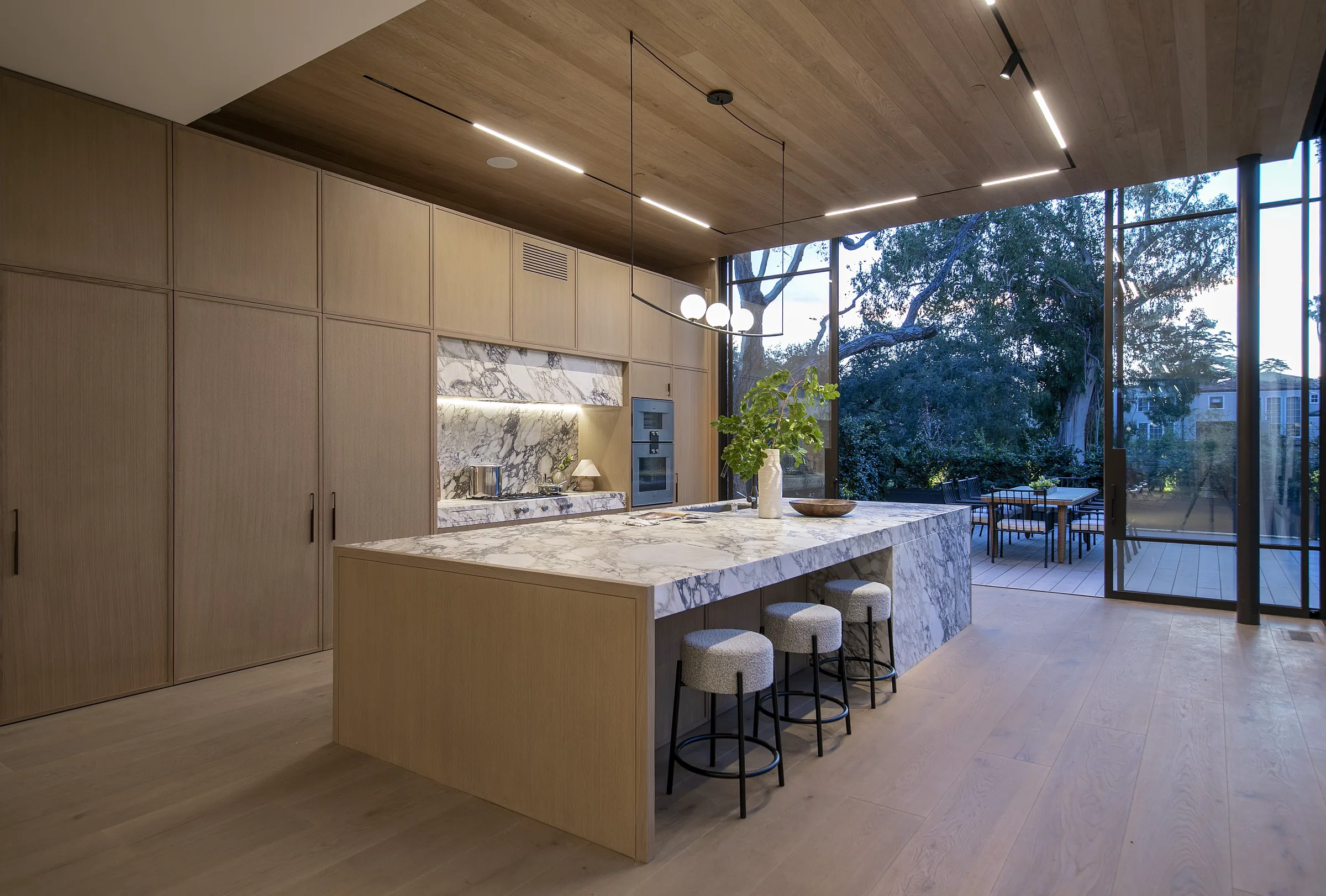 Open-plan kitchen with a monolithic Viola marble waterfall island in a white-and-plum veined field, paired with full-height white-oak cabinetry and a matching Viola marble cooktop backsplash niche, bouclé counter stools, a slim globe pendant, and floor-to-ceiling steel-framed glass opening to a wooded garden terrace.