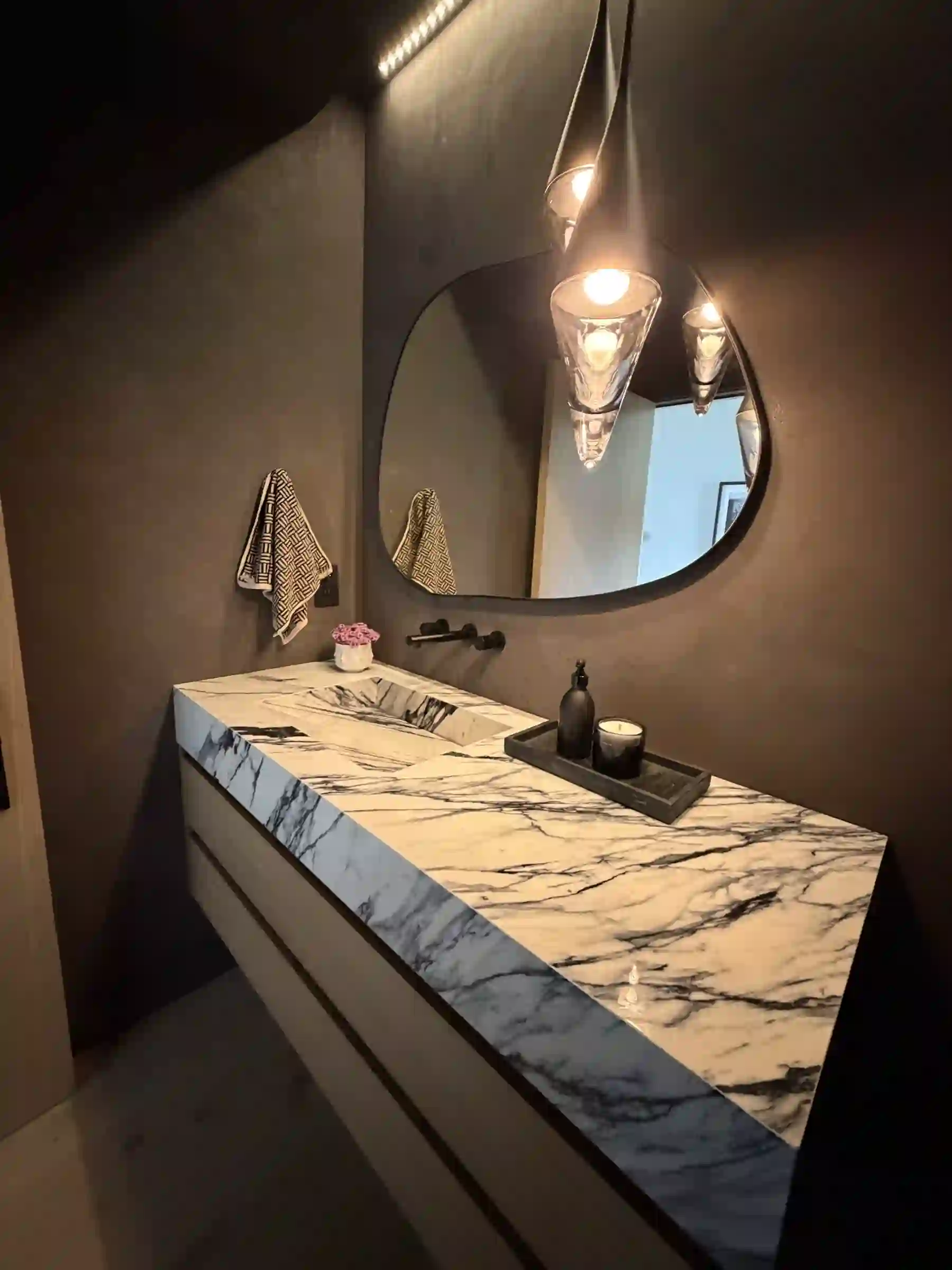 Close view of a Calacatta Viola marble powder-room vanity — the veined marble top and carved integral sink with a wall-mounted matte-black faucet, a round black-framed mirror, and a glowing glass pendant against dark plaster walls.