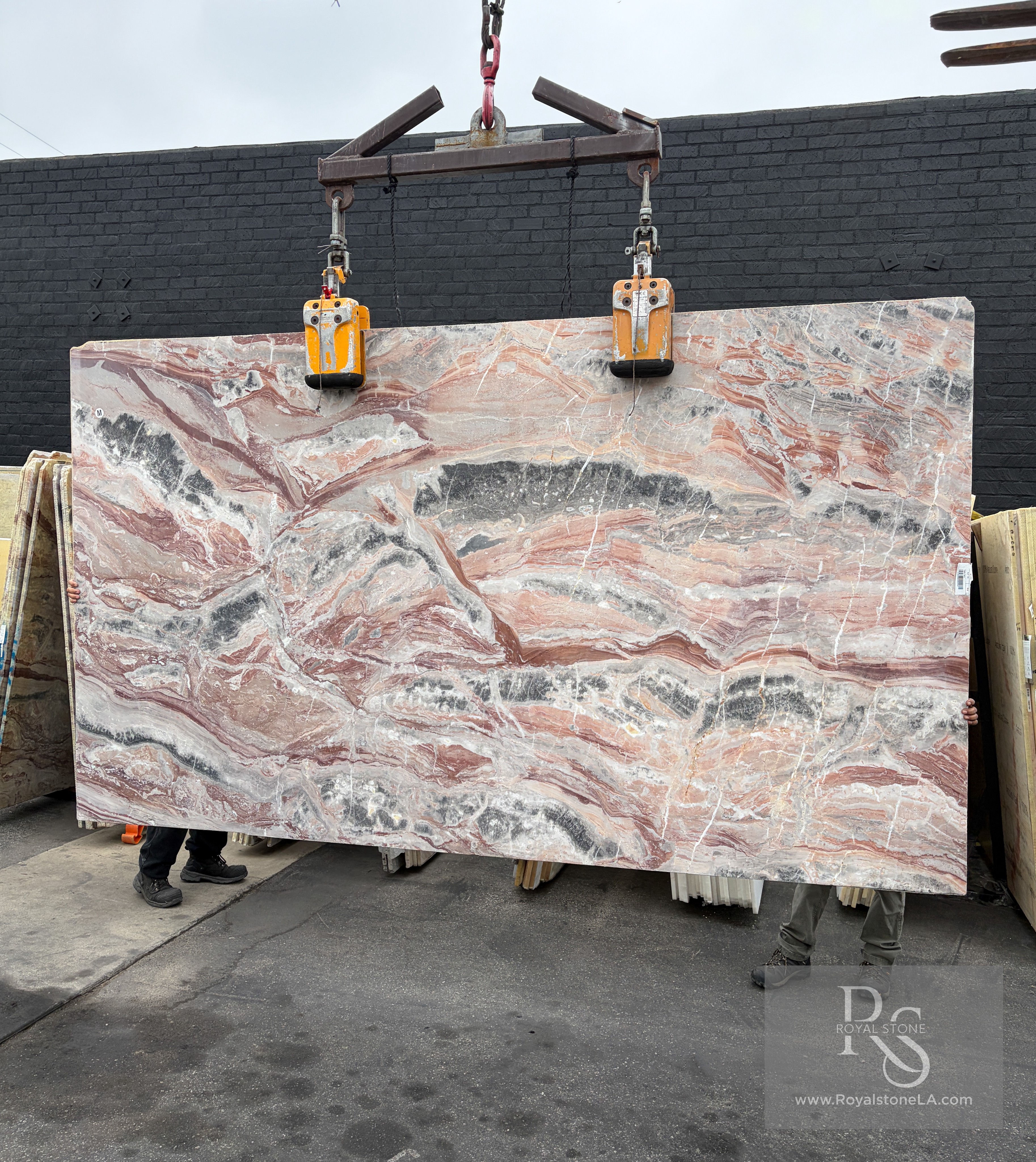 Viola Arabescato Orobico Rosso honed Italian marble slab, 75 by 130 inches, with horizontal rose, coral, charcoal, and white quartz currents, suspended from a crane in front of the Royal Stone yard wall